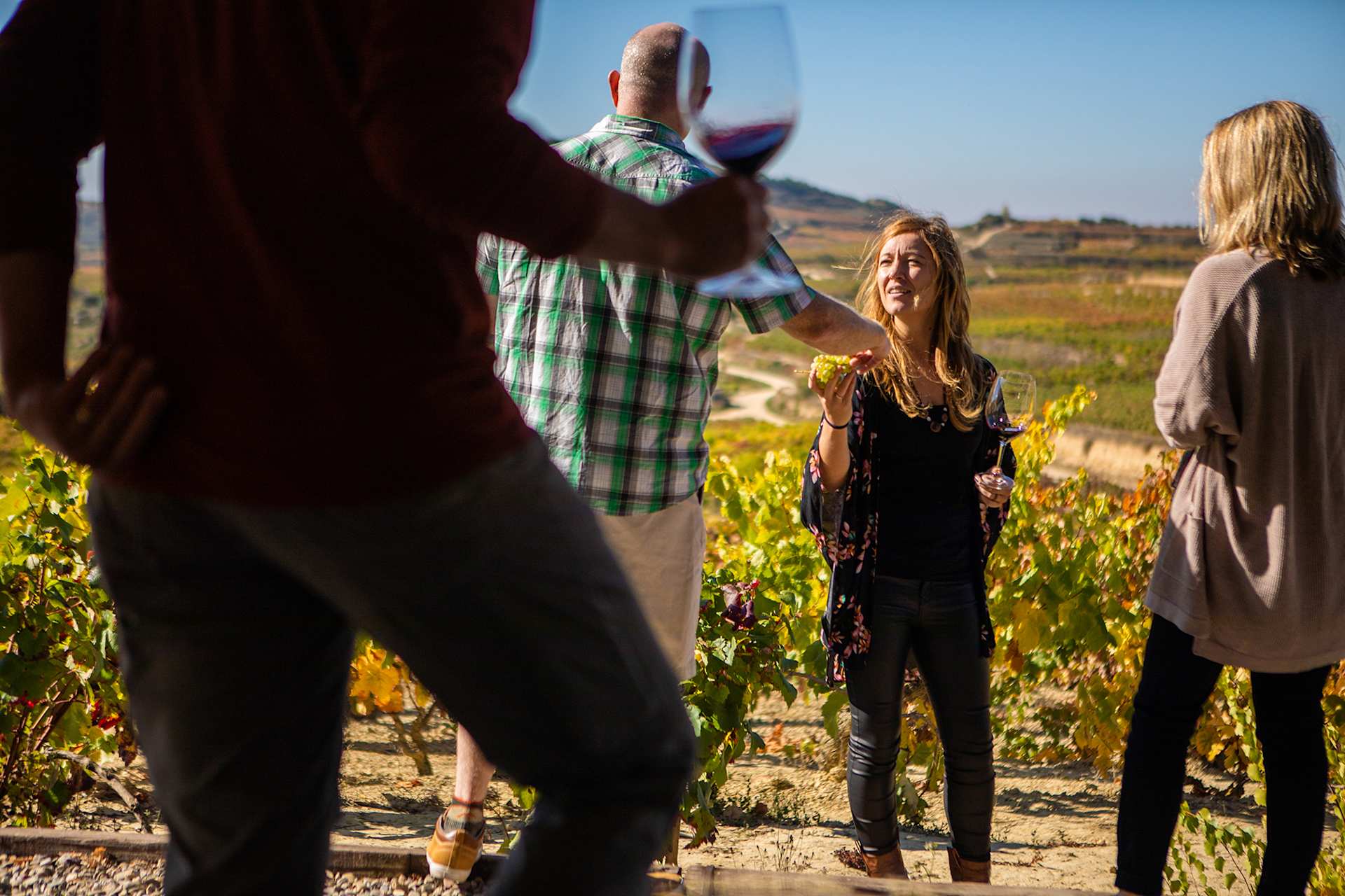 A group of people, including a woman in a floral dress, standing in a vineyard with mountains in the background.