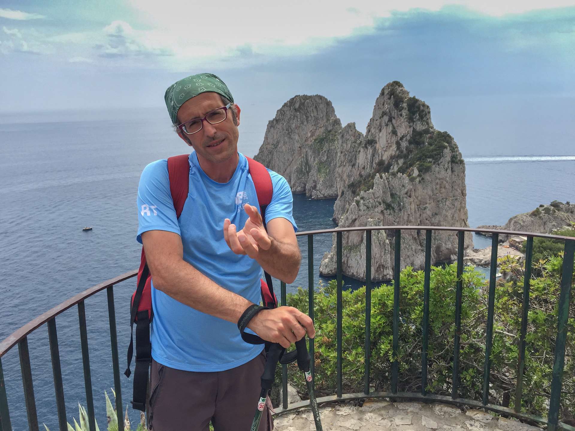 A man wearing a blue shirt, red backpack, and a green cap stands on a balcony overlooking a scenic coastal landscape with towering rock formations in the background.