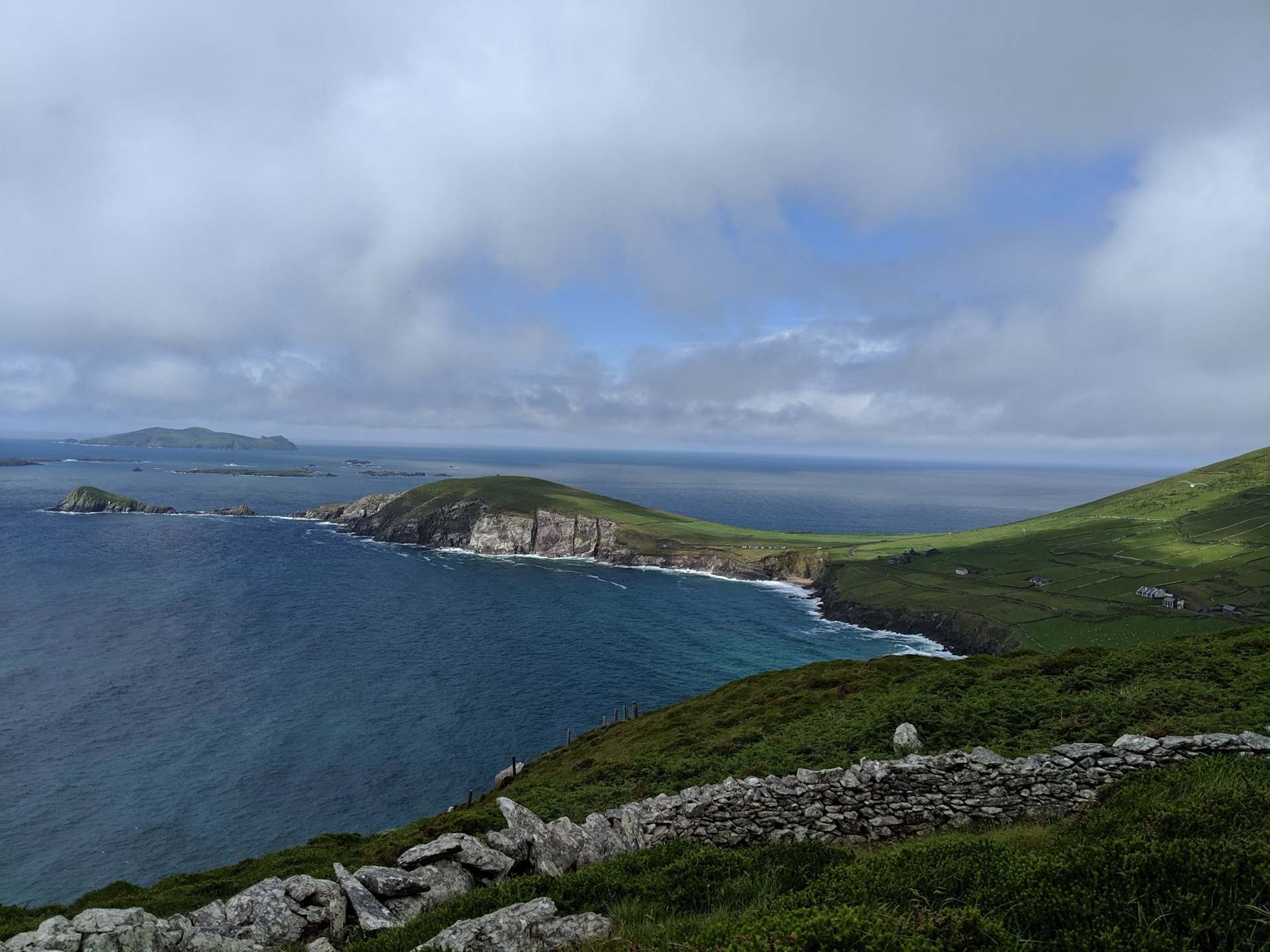 A rugged, green coastline with rocky cliffs overlooking a vast, blue ocean under a cloudy, overcast sky.