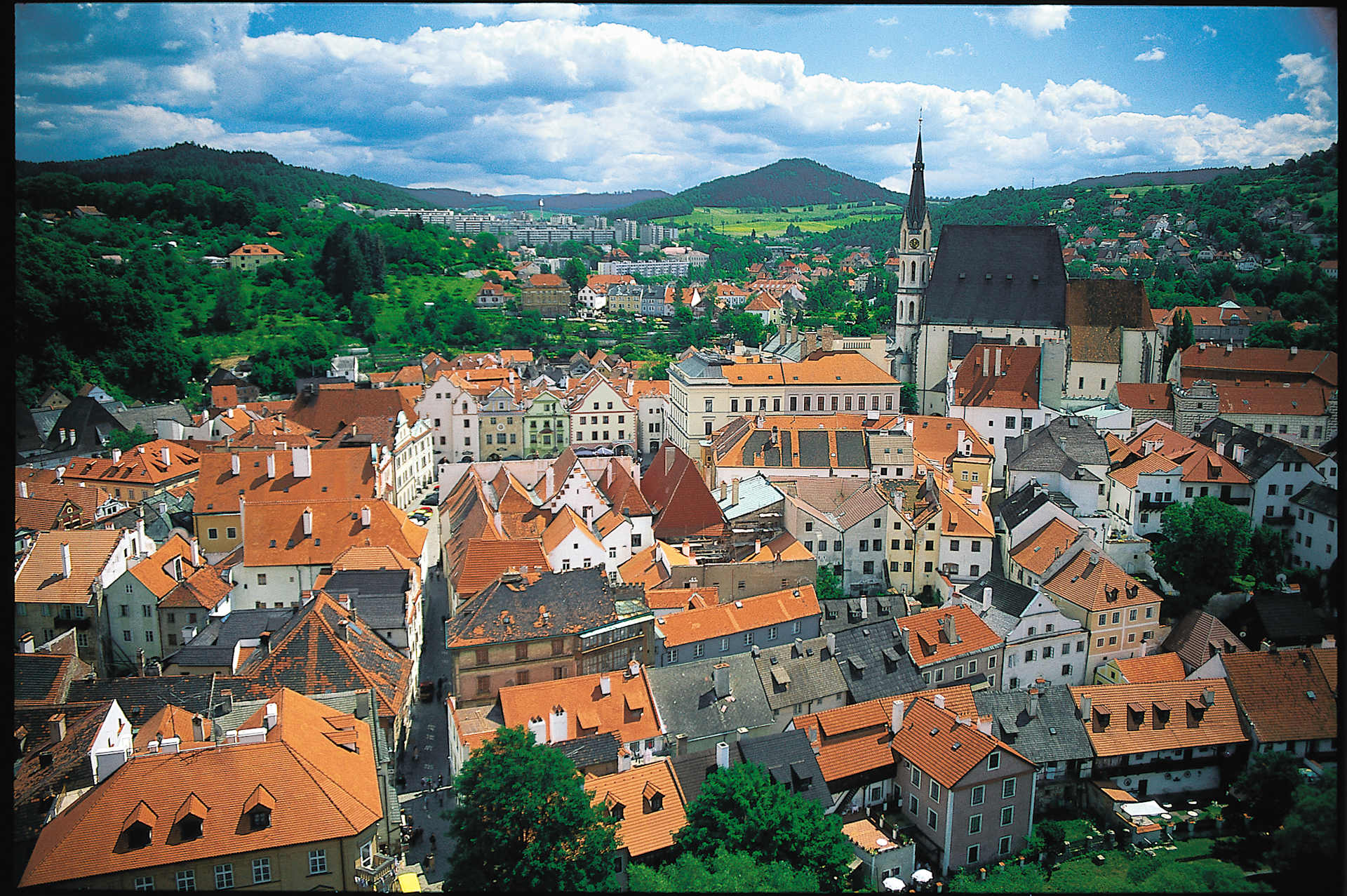 A picturesque European city nestled in a valley, with colorful rooftops, church spires, and lush green hills in the background under a cloudy blue sky.
