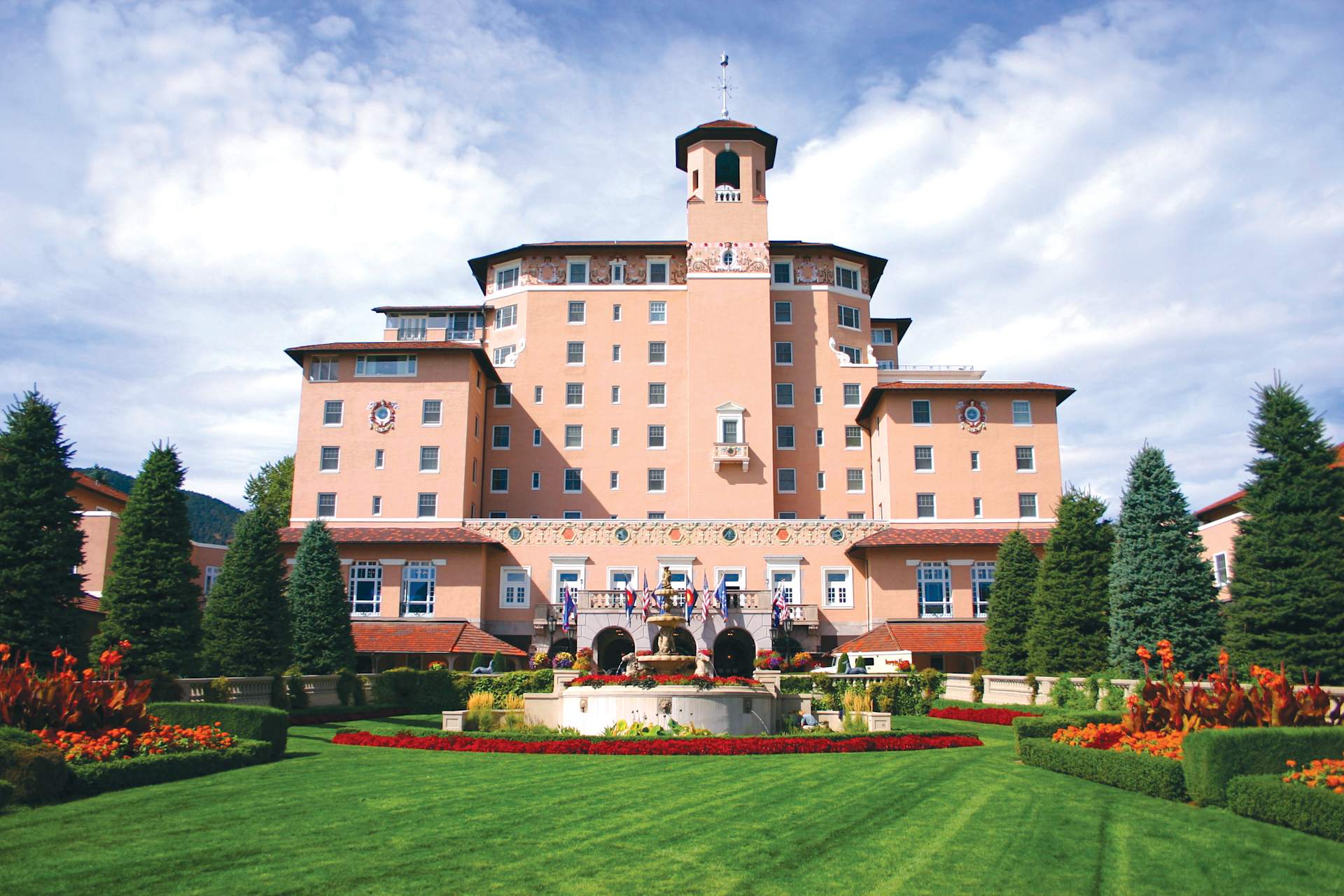 A grand, ornate hotel with a pink facade stands in a well-manicured garden, surrounded by lush greenery and vibrant flower beds in the foreground.