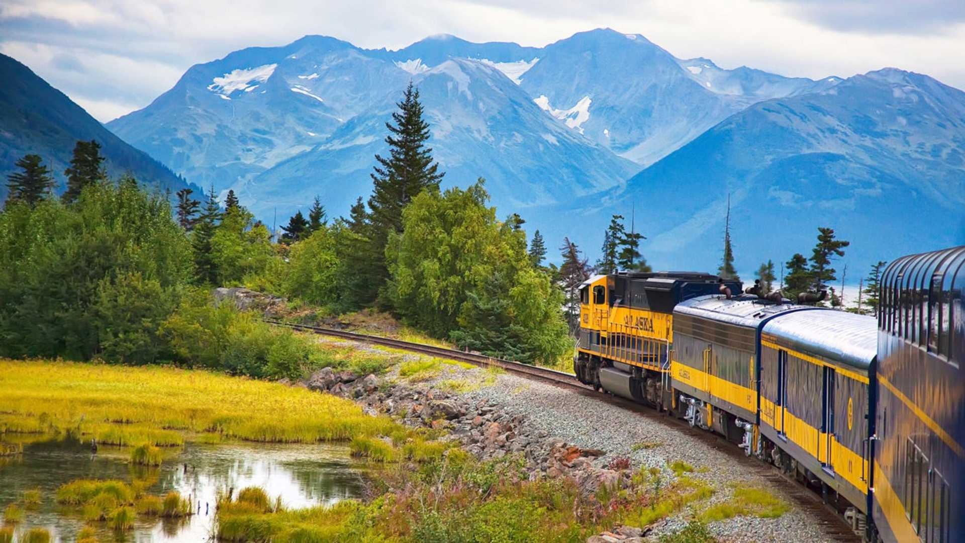 A scenic train journey through a lush, mountainous landscape, with a yellow locomotive traversing the tracks alongside a tranquil pond and surrounded by towering, snow-capped peaks.