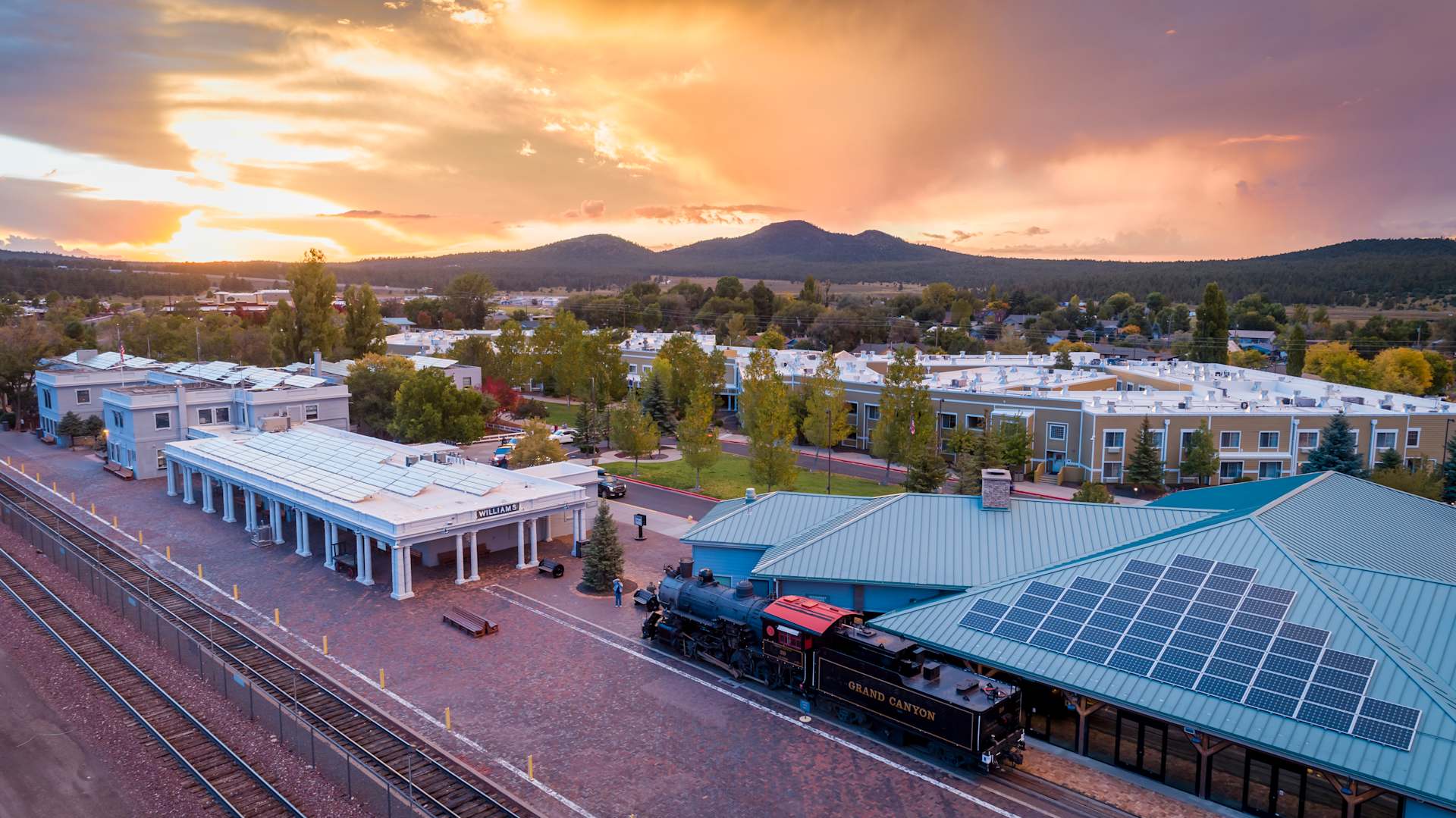 A vibrant sunset casts a warm glow over a picturesque town nestled among rolling hills, with a historic steam locomotive passing through the foreground.