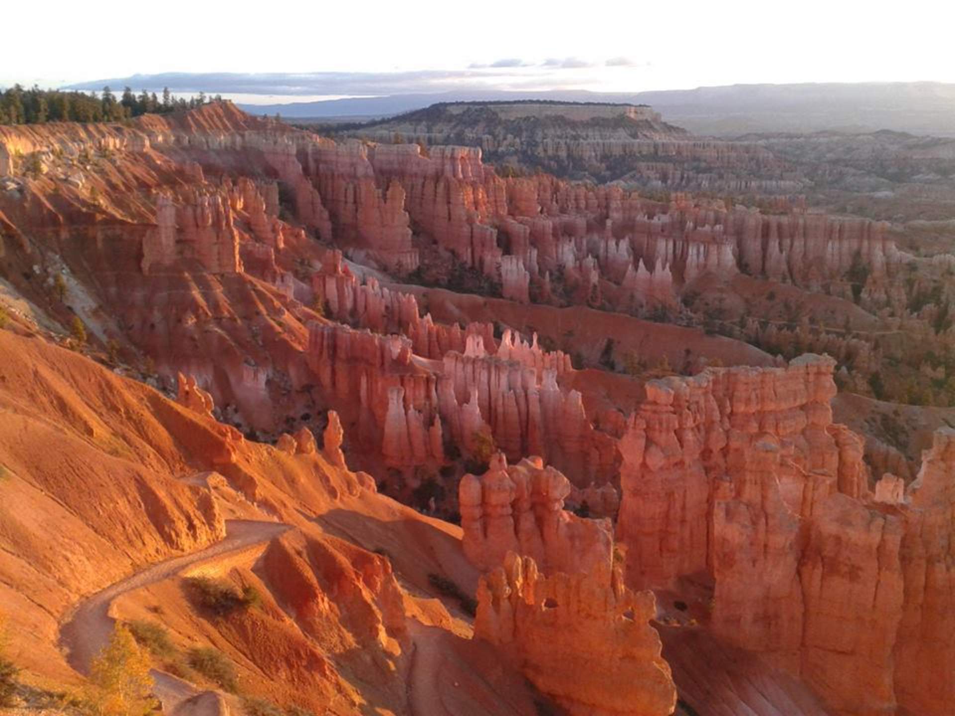 A vast, rugged landscape of towering red rock formations and deep canyons, set against a hazy, distant horizon under a cloudy sky.