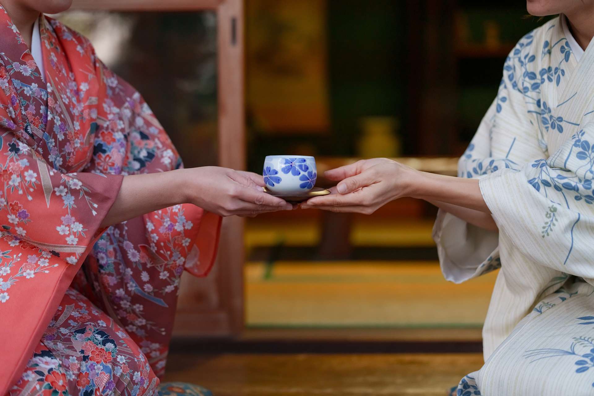 Two individuals in traditional Japanese attire, one wearing a vibrant red kimono with floral patterns and the other in a white kimono with blue floral designs, are exchanging a small blue-and-white ceramic cup in what appears to be a traditional Japanese setting.
