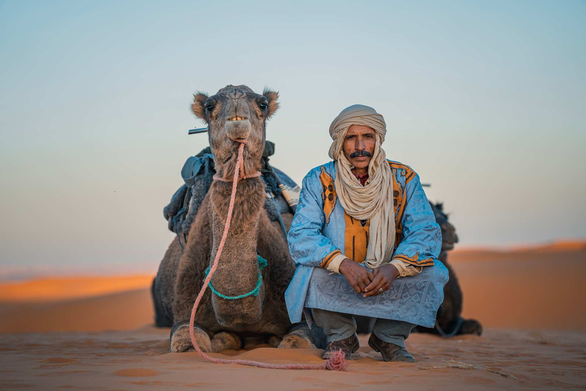 Two men, one riding a camel, stand in a desert landscape with a vibrant orange sky in the background.