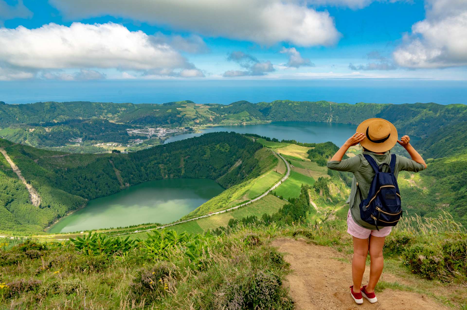 A hiker with a backpack and hat stands on a grassy overlook, gazing at a vast caldera with two lakes and a distant ocean under a cloudy blue sky.