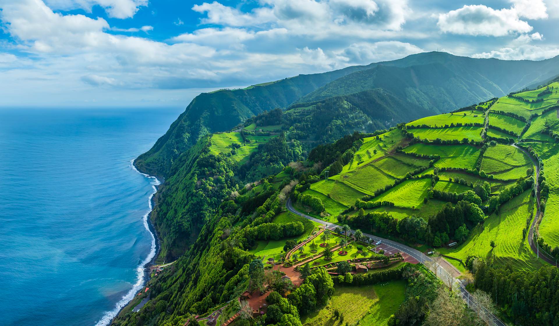 Lush green terraced fields and a winding road lead down to the deep blue ocean along a dramatic coastline.