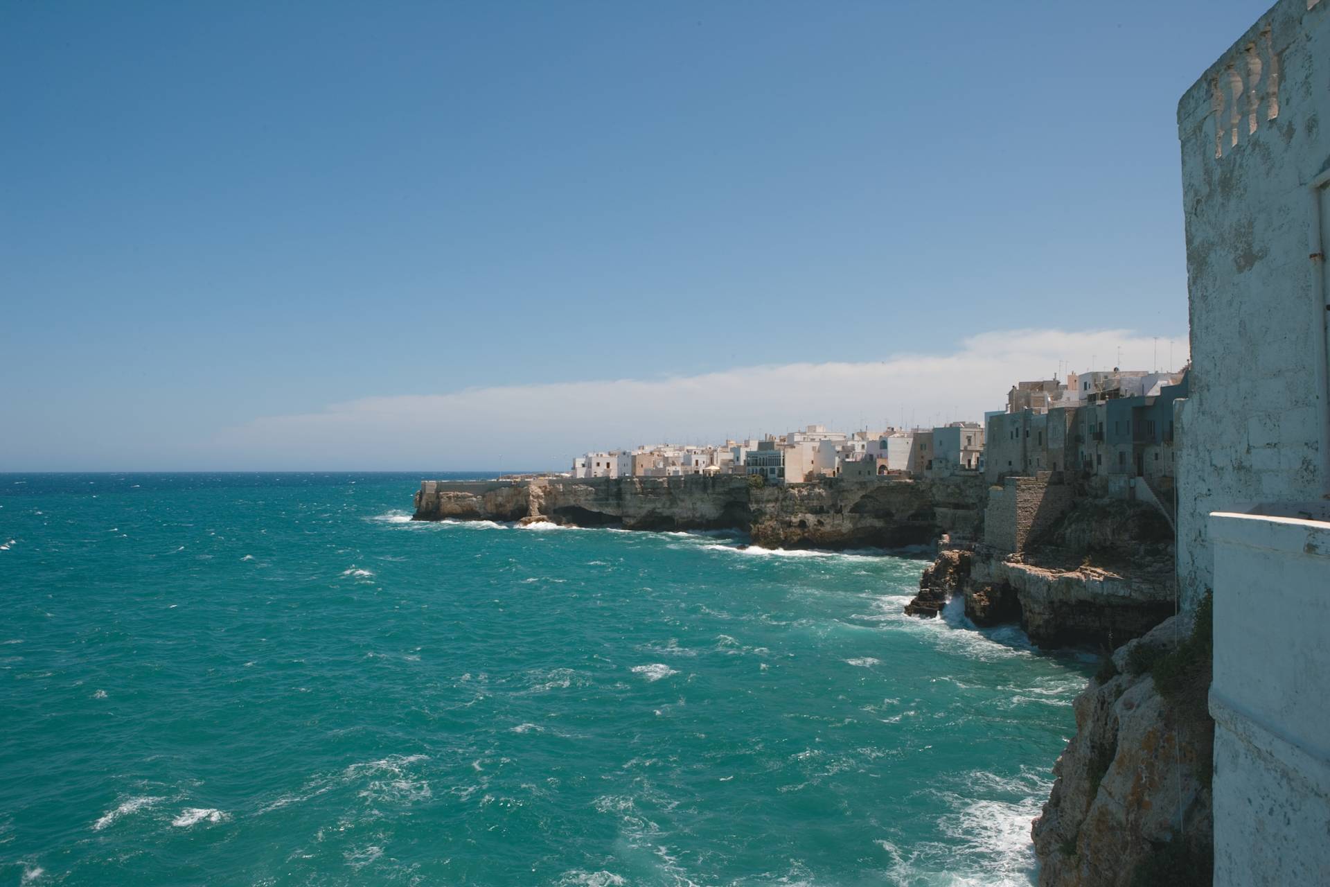 Turquoise waves crash against rocky cliffs in the foreground, leading to a white-washed town perched on the coastline under a clear blue sky in the background.