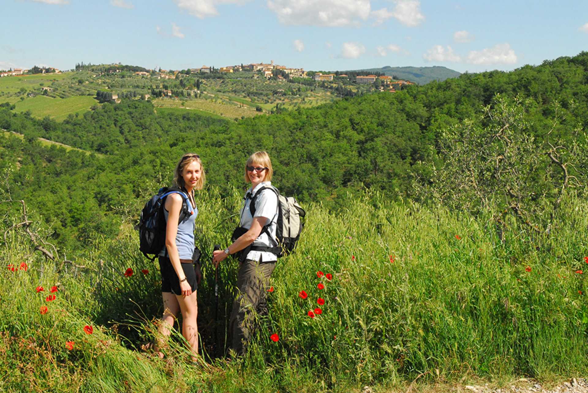 Two women with backpacks stand in a field of tall green grass and red poppies, with rolling green hills and a distant town under a blue sky in the background.