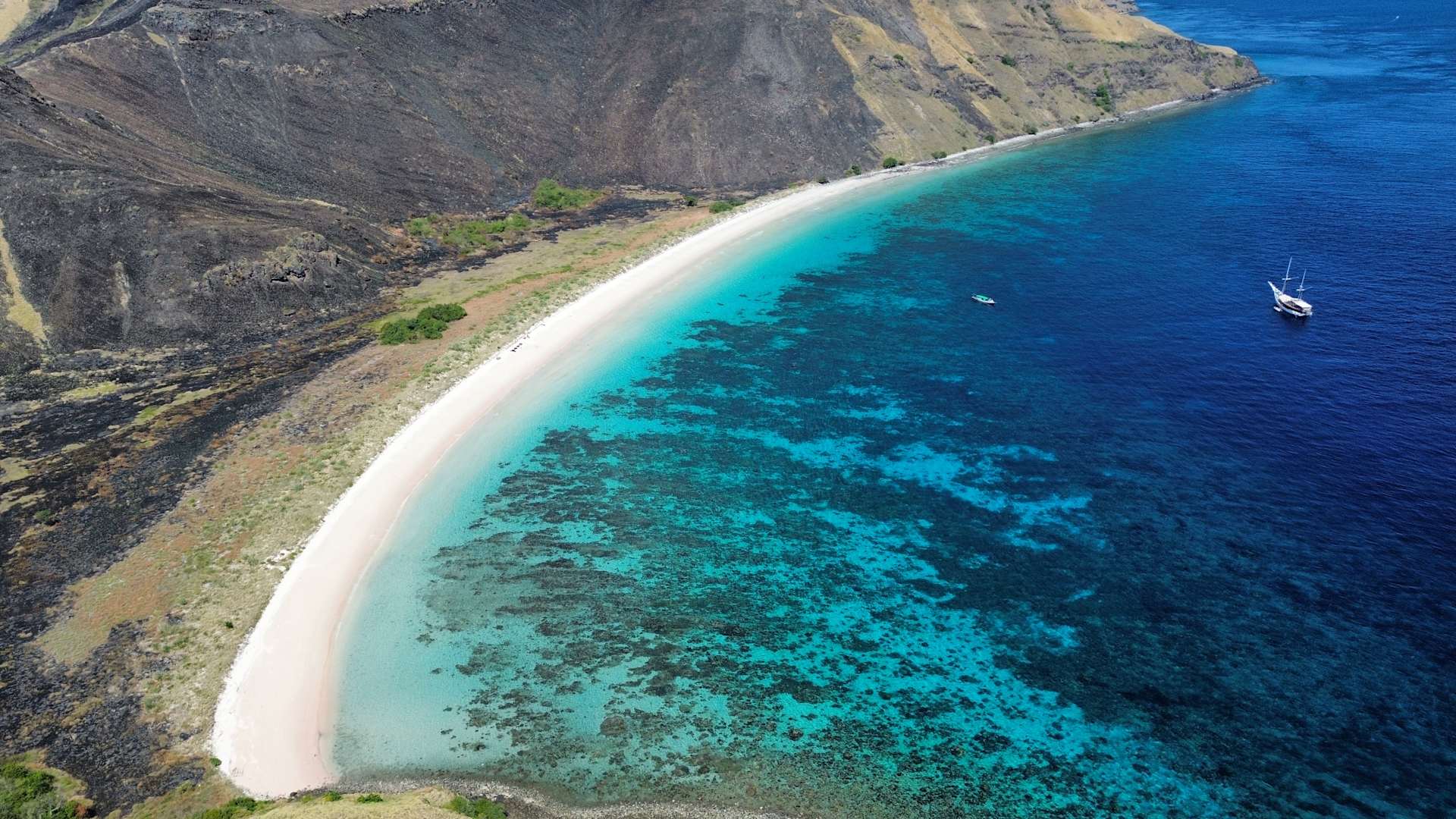 A white sandy beach meets turquoise waters with a white boat in the foreground, set against a backdrop of rugged, brown mountains.