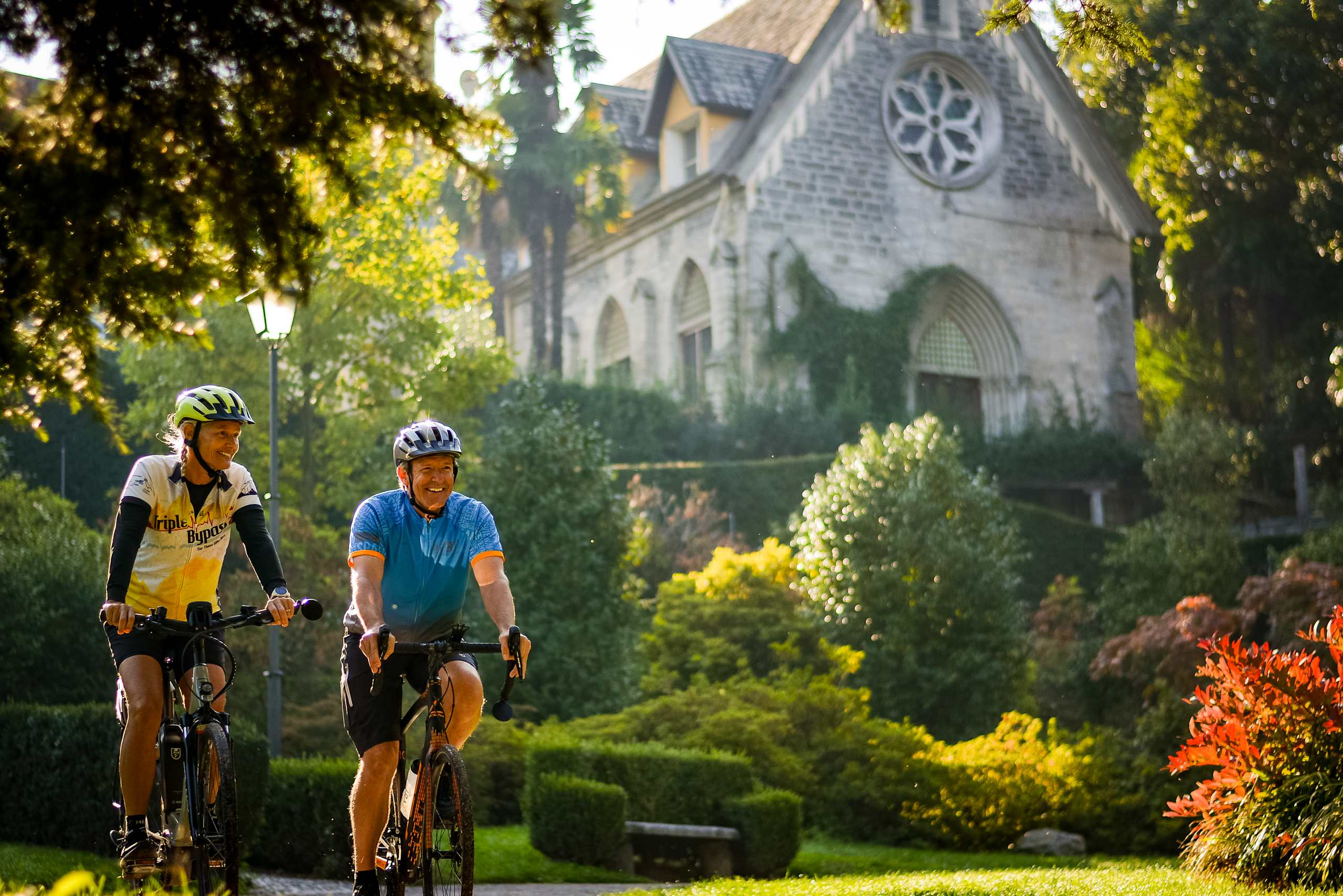 Two cyclists riding their bicycles on a path surrounded by lush greenery and a picturesque church in the background.