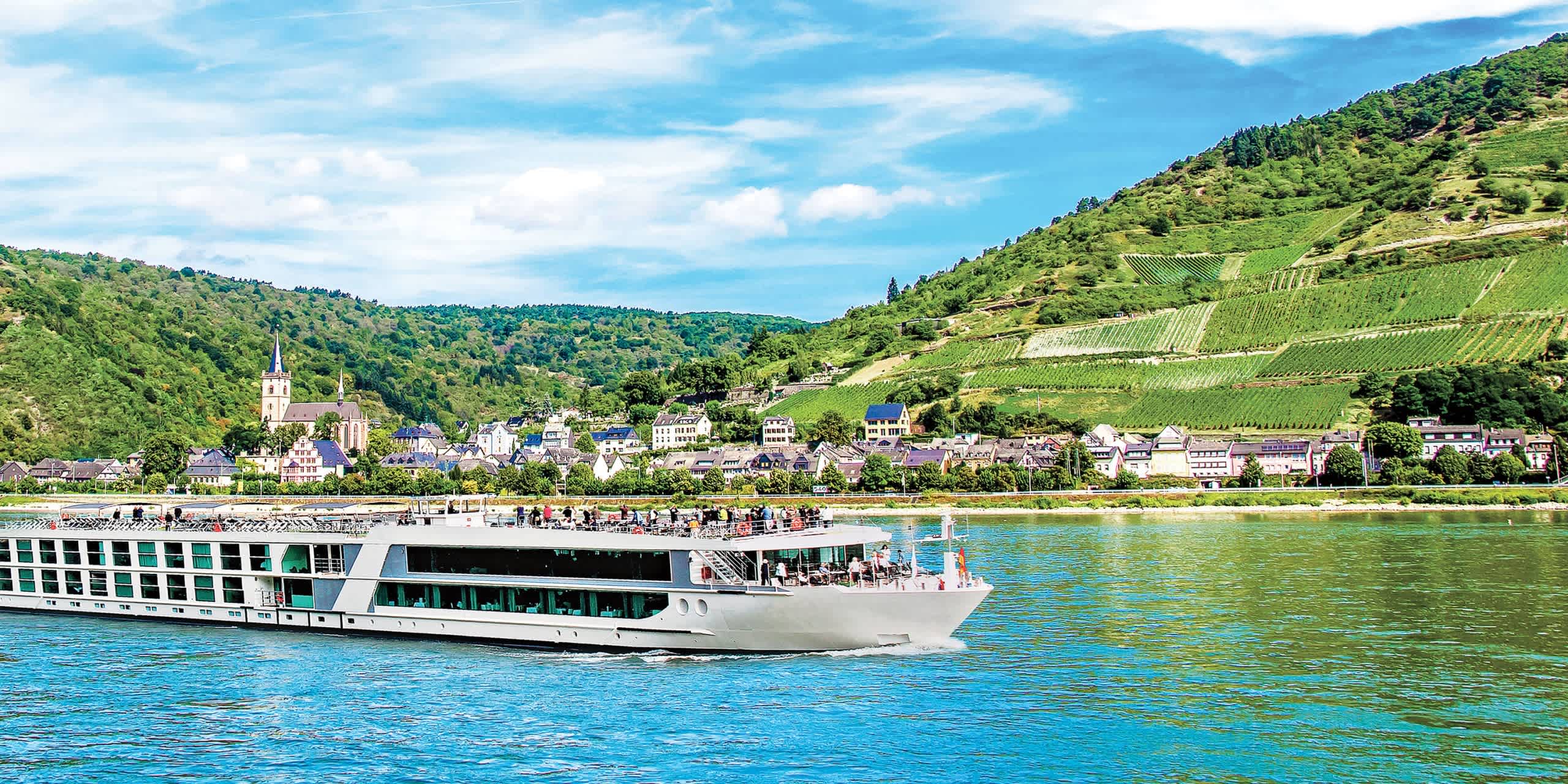 A scenic river cruise ship sails along a picturesque river surrounded by lush, verdant hills and a charming town nestled in the valley.