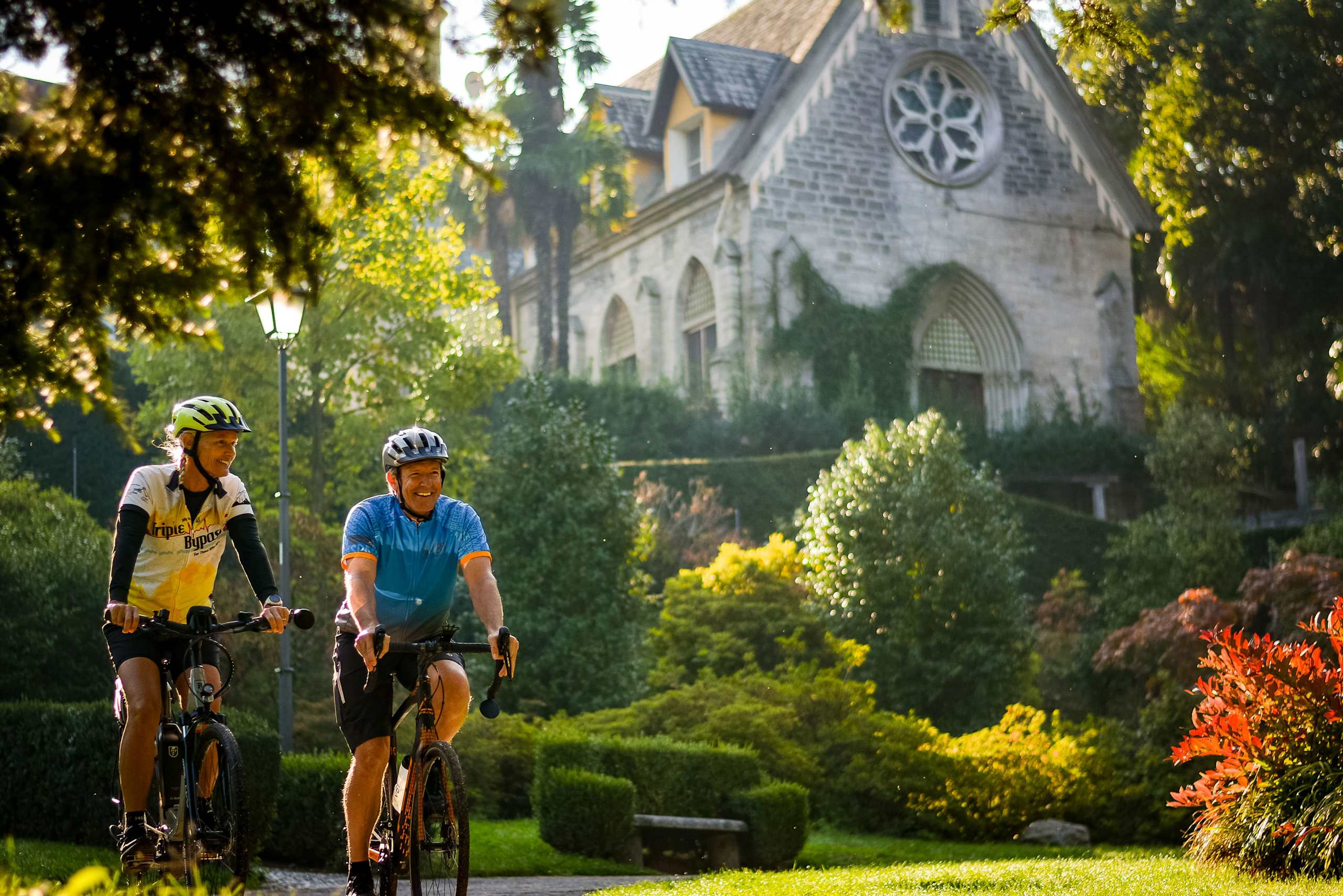 Two cyclists riding their bicycles through a lush, verdant garden with a grand, gothic-style church or cathedral in the background.