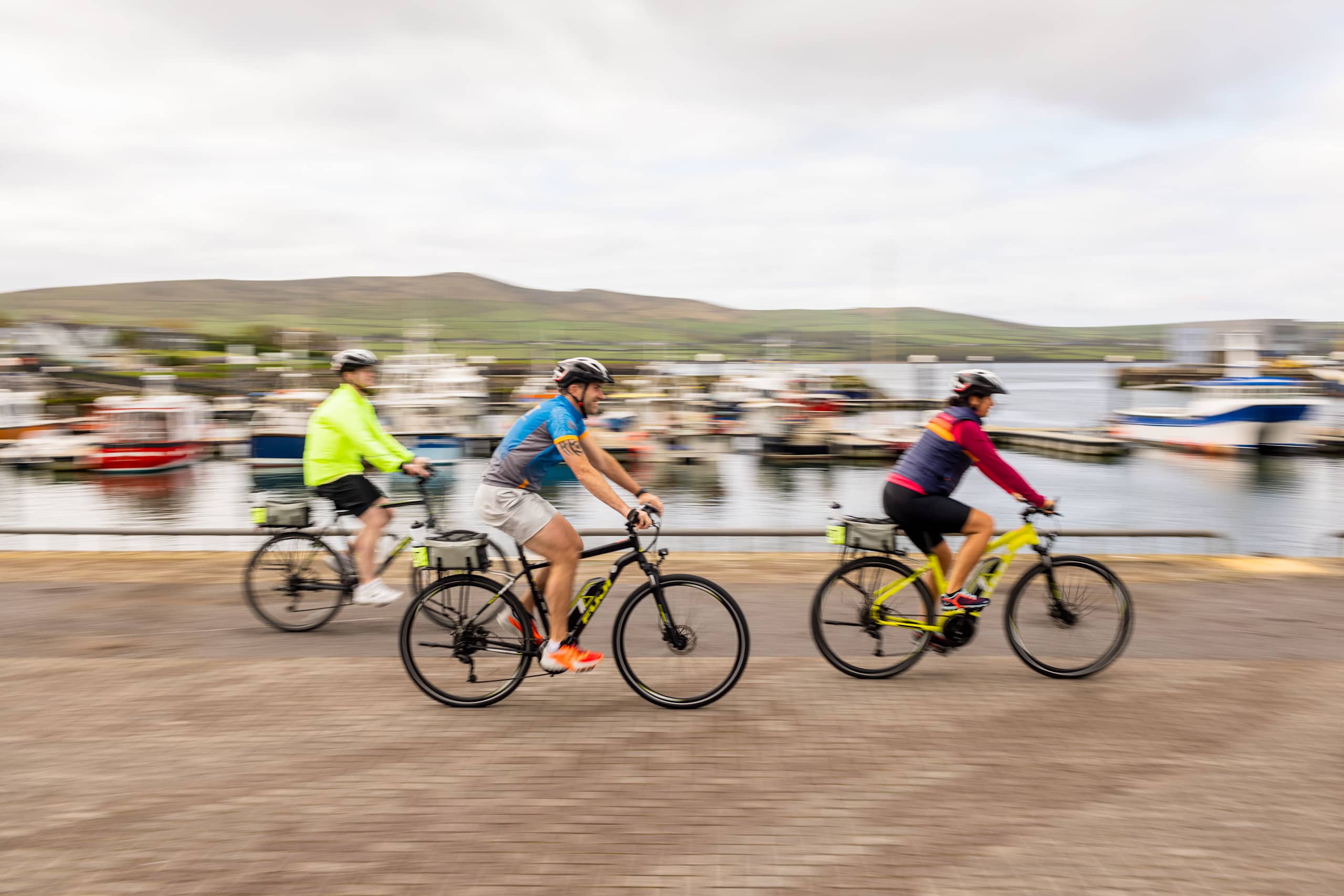 A group of cyclists riding on bicycles in a coastal town, with mountains and buildings visible in the background.