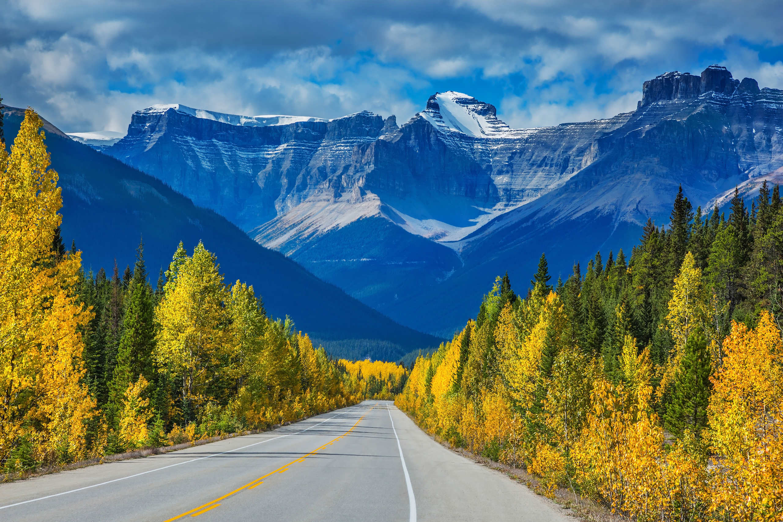 A winding road leads through a vibrant autumn landscape, with towering snow-capped mountains rising in the background against a dramatic cloudy sky.