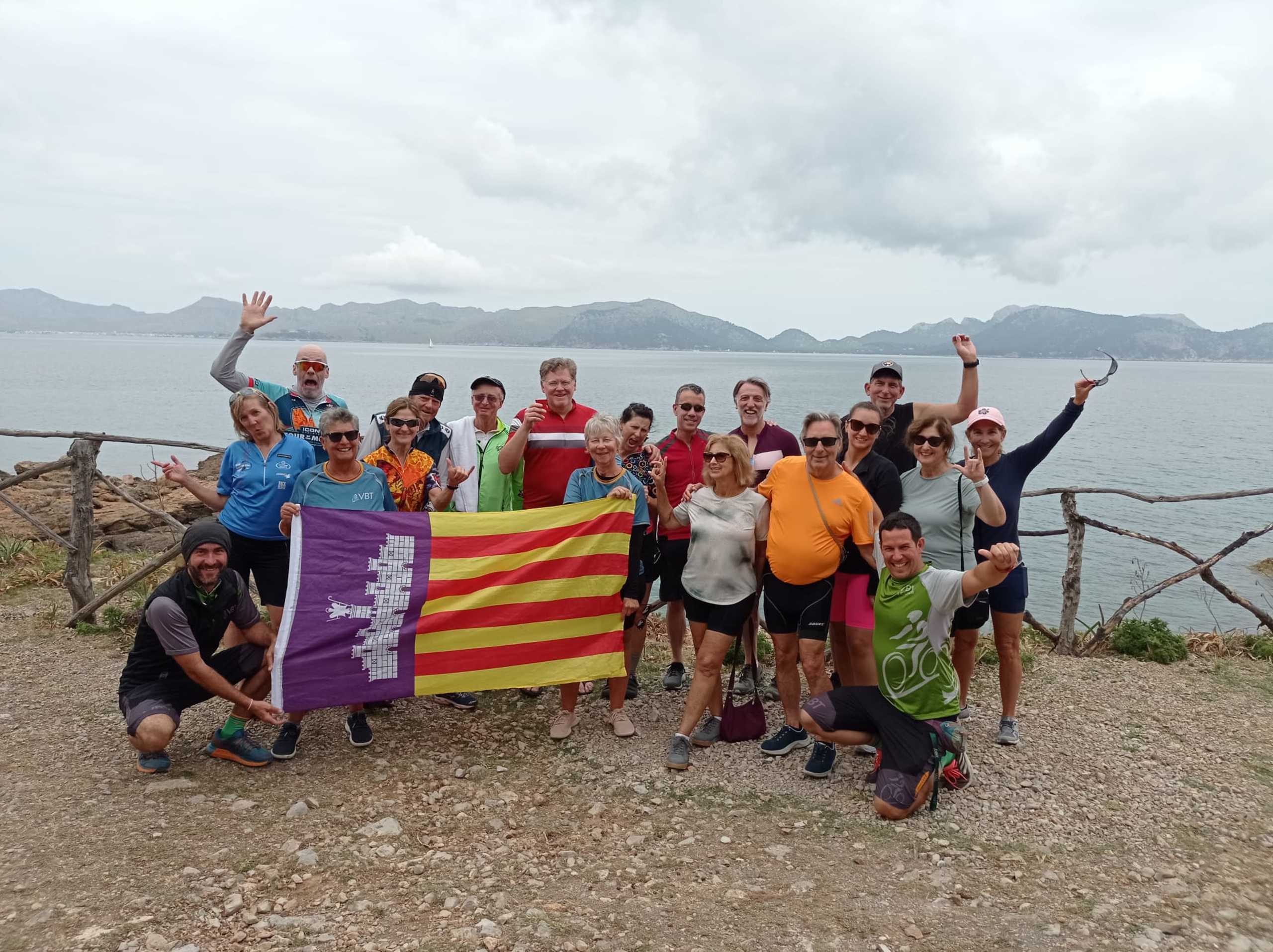 A group of people, some wearing colorful clothing, standing together on a rocky shore with mountains in the background, holding up a flag.