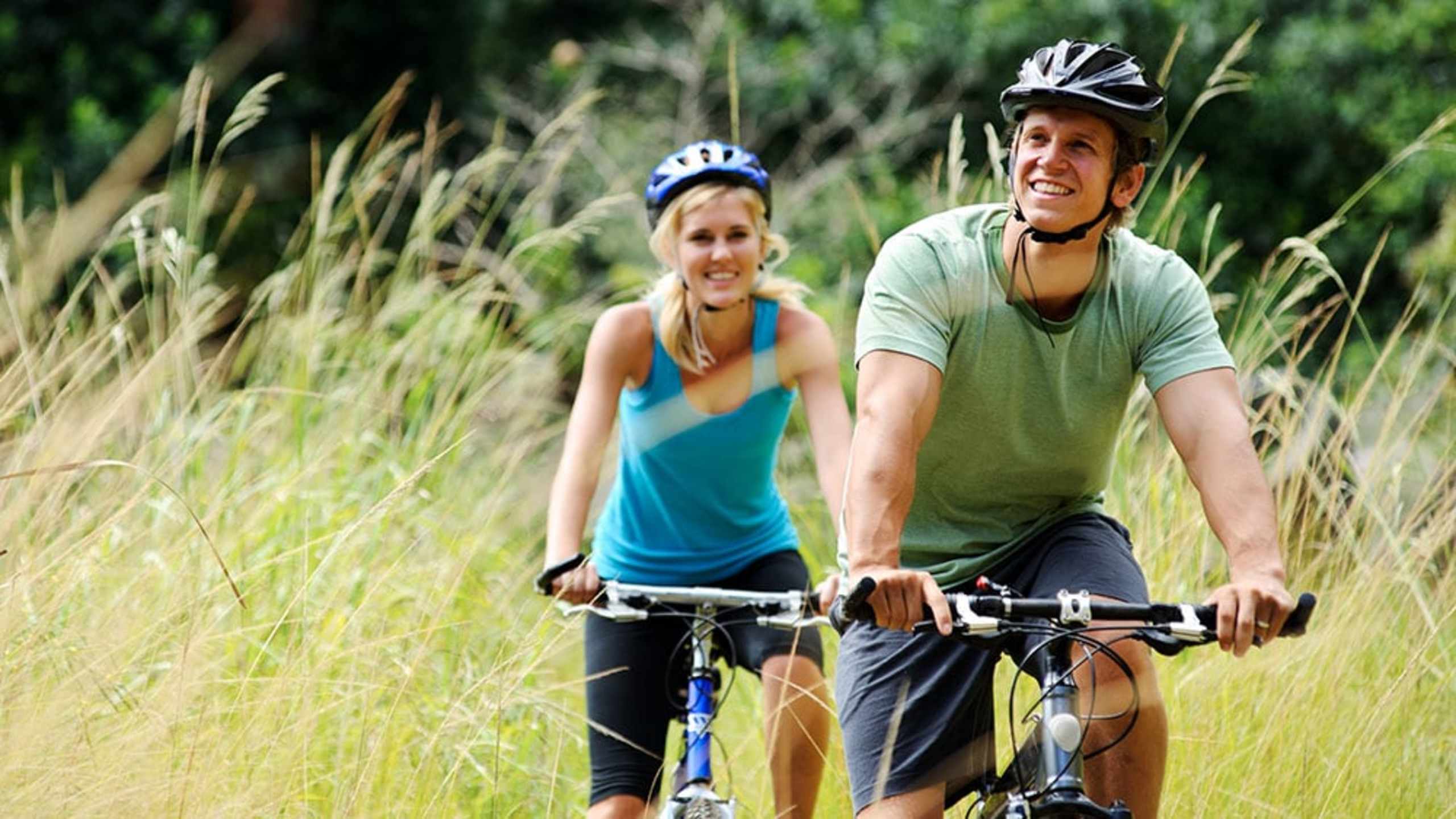 Two people, a man and a woman, are riding bicycles through a grassy field with tall vegetation in the background.