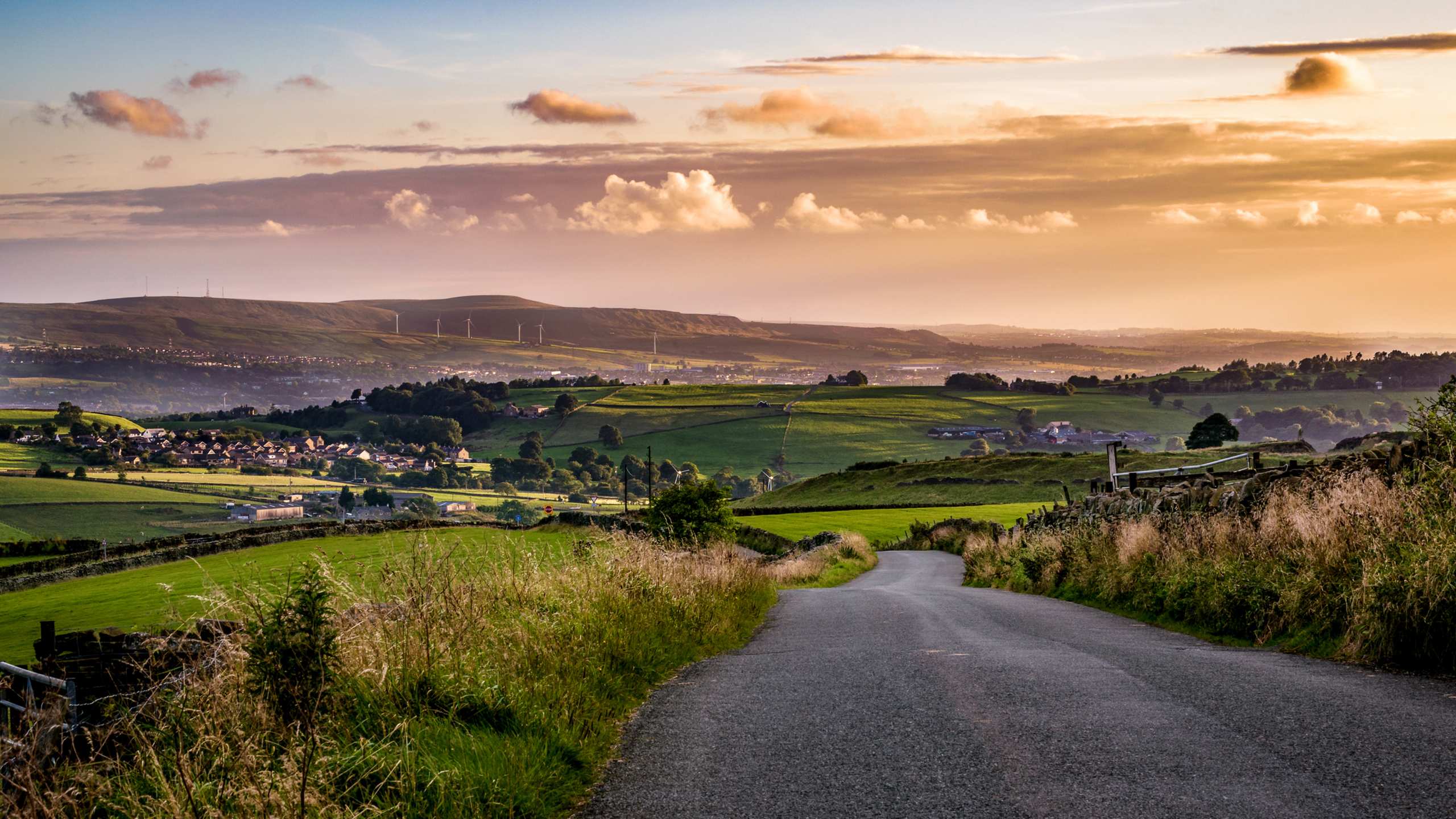 A winding dirt road leads through a lush, rolling countryside landscape, with distant hills and a dramatic sky filled with clouds at sunset.