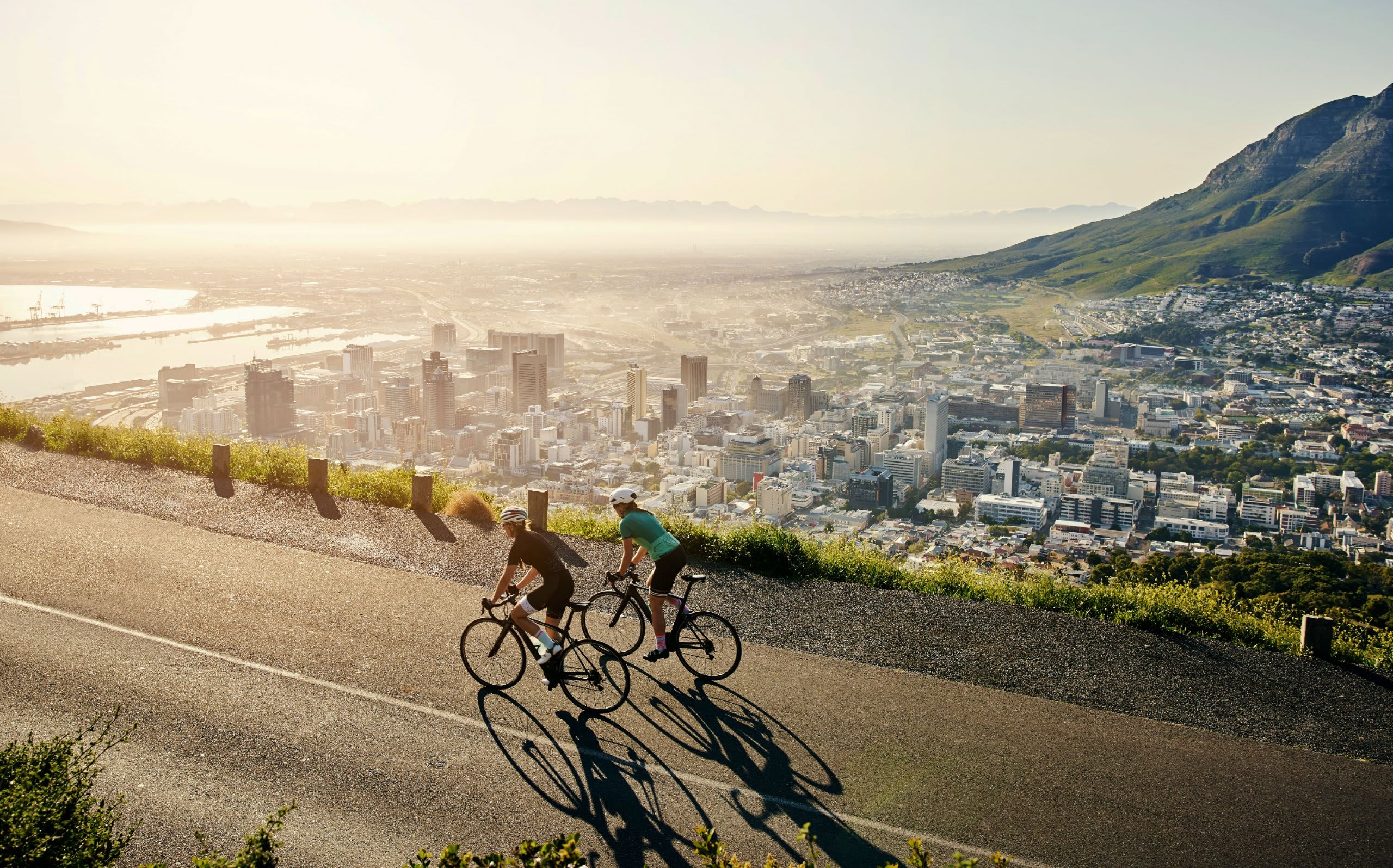 Two cyclists riding bicycles on a winding road with a stunning cityscape and mountainous landscape in the background.