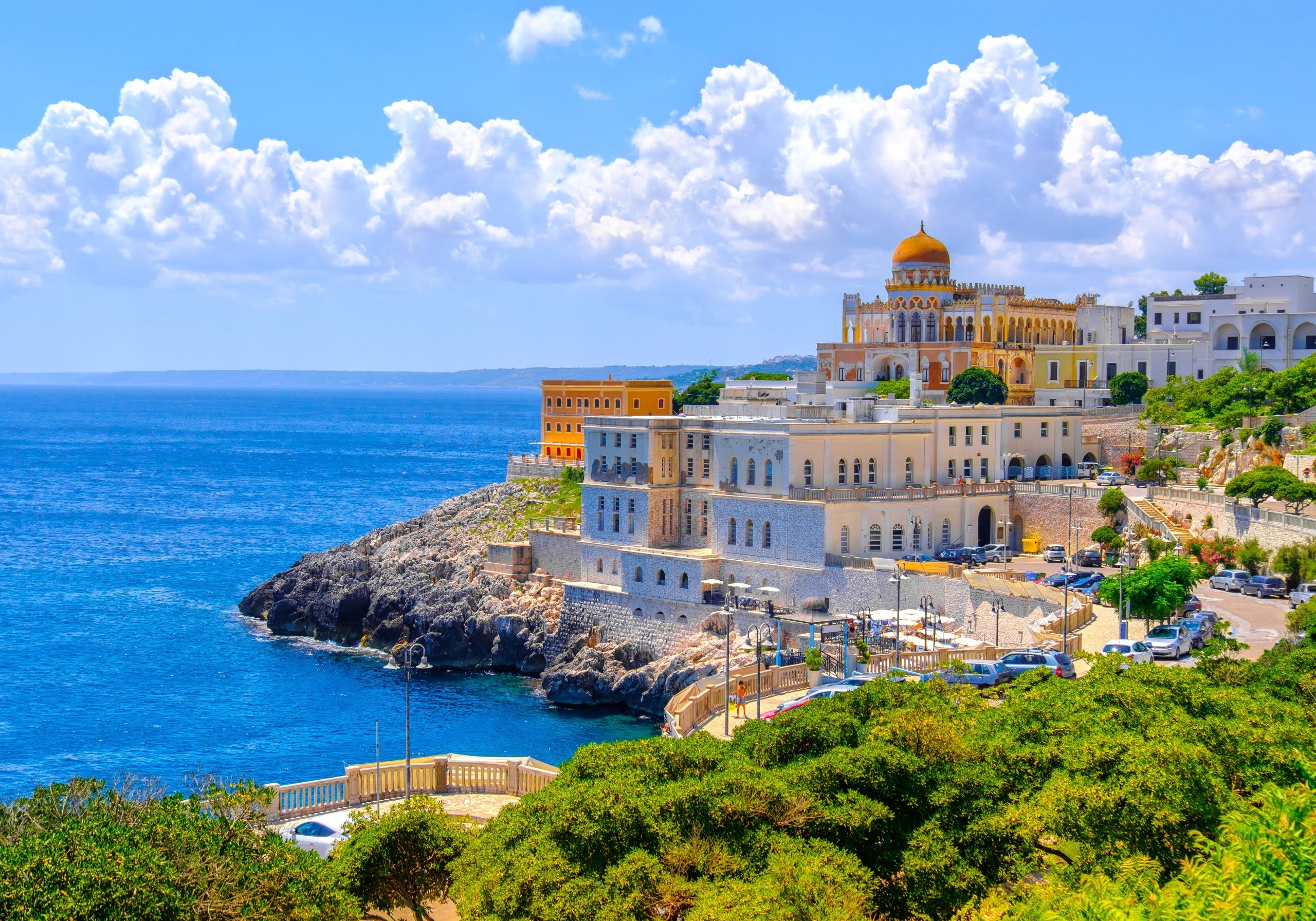 A picturesque coastal town with colorful buildings nestled on a cliff overlooking the vibrant blue waters of the sea, set against a backdrop of fluffy white clouds in a bright, clear sky.