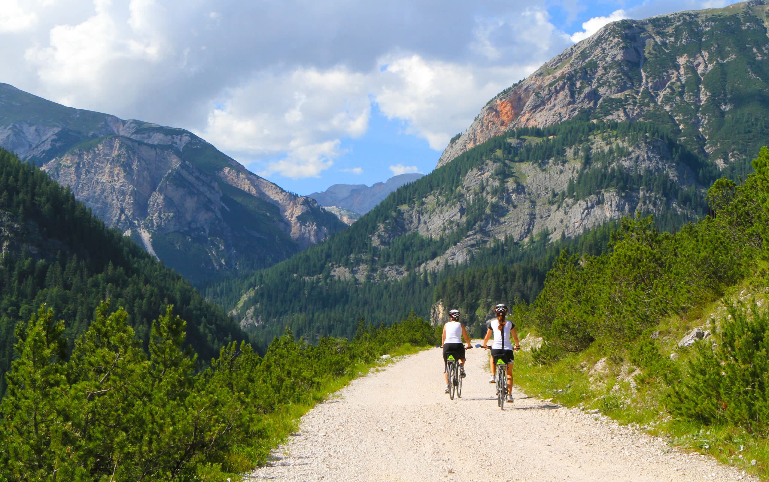 Two cyclists riding on a dirt path through a lush, mountainous landscape with towering peaks in the background.
