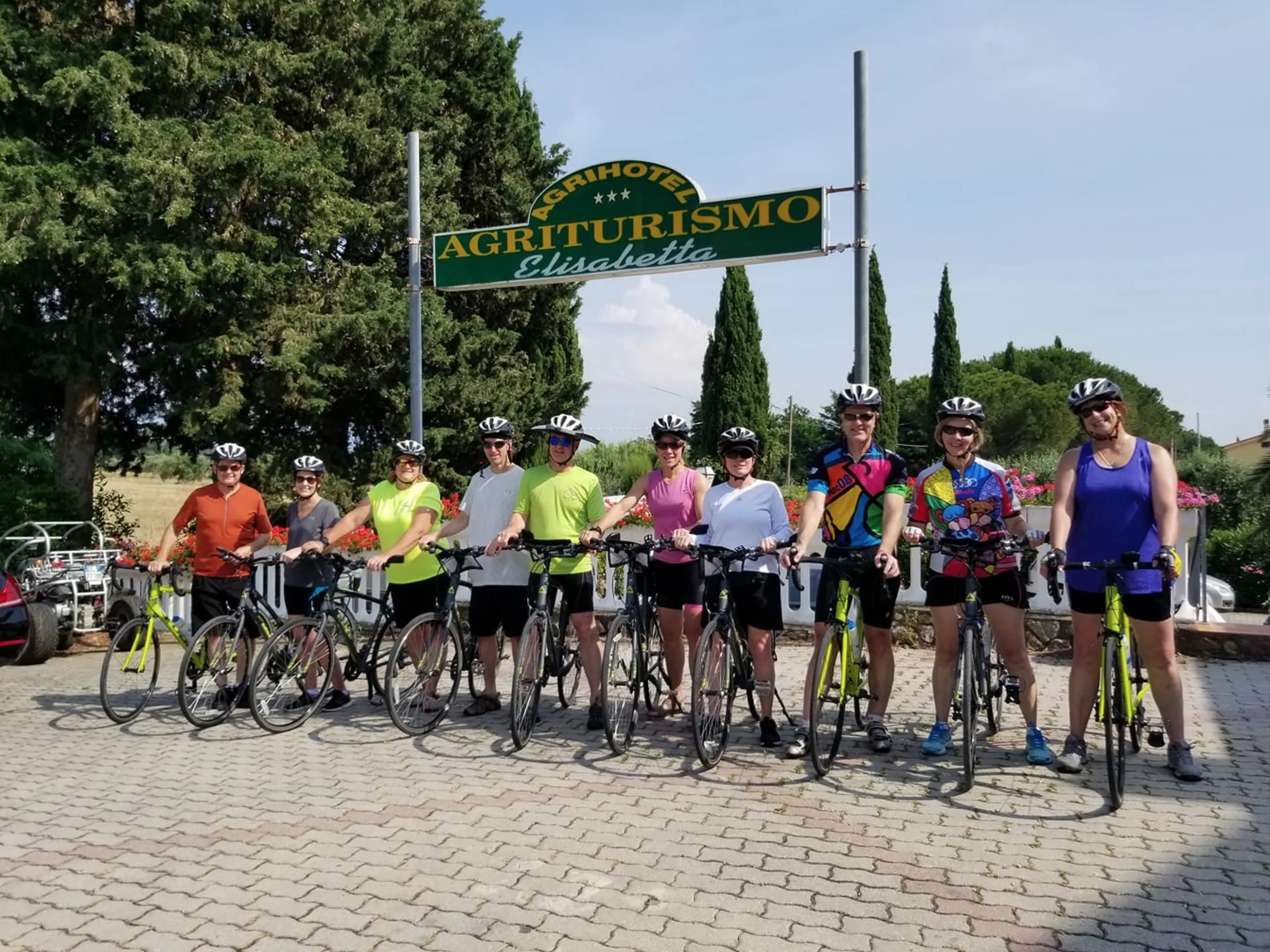 A group of cyclists in colorful cycling gear standing in front of a sign for "Agriturisimo", with a backdrop of trees and a clear sky.