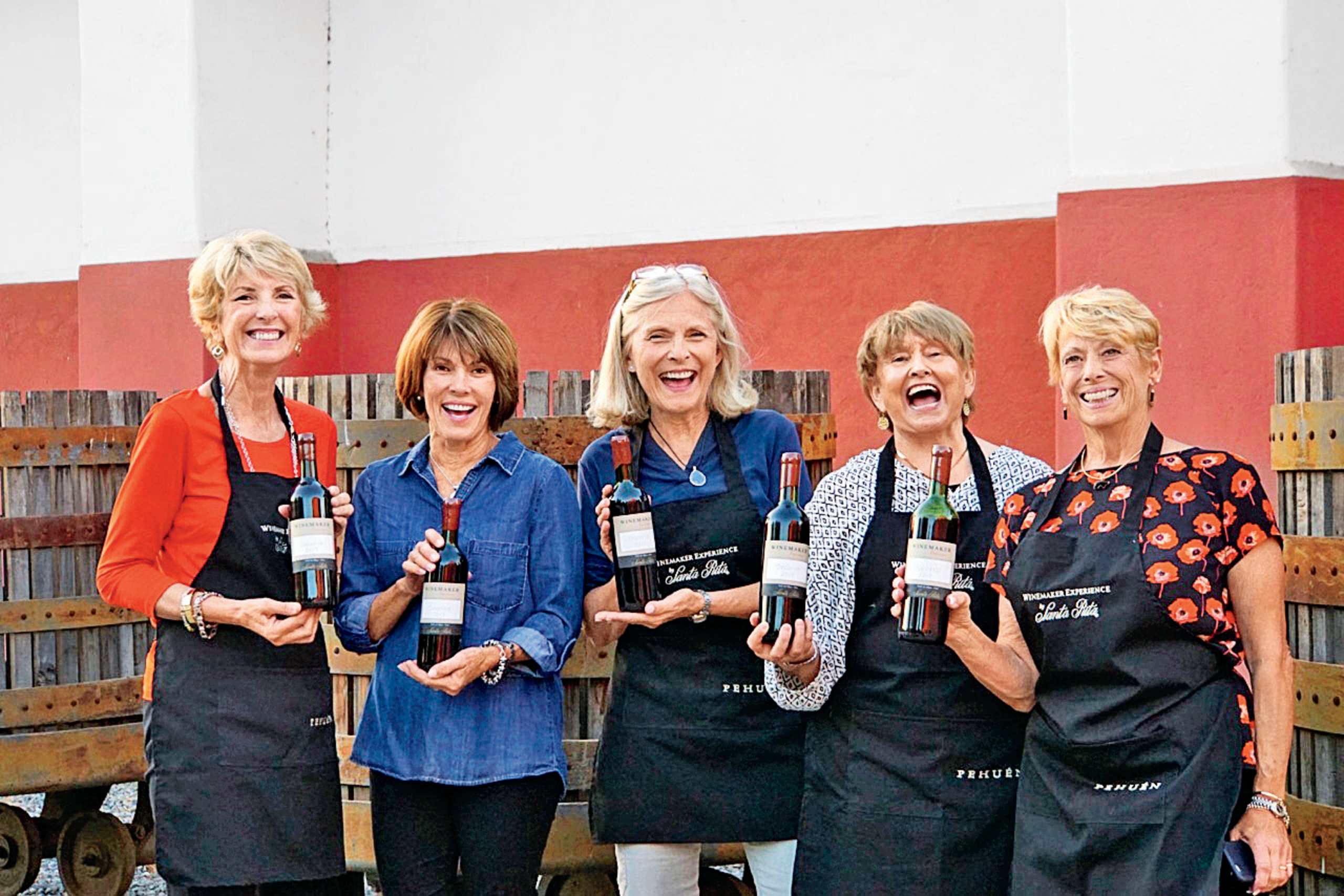 A group of five smiling women in aprons and casual clothing stand in front of a wall of wine bottles, appearing to be employees or staff of a winery or wine shop.