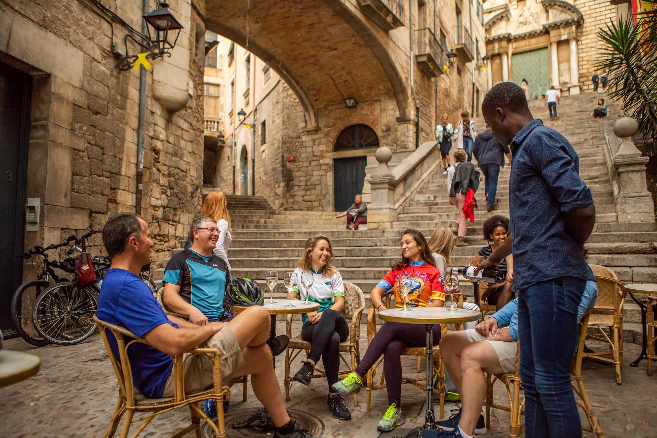 A group of people, both men and women, are sitting at outdoor tables in a historic European-style alleyway, surrounded by old stone buildings and arches.