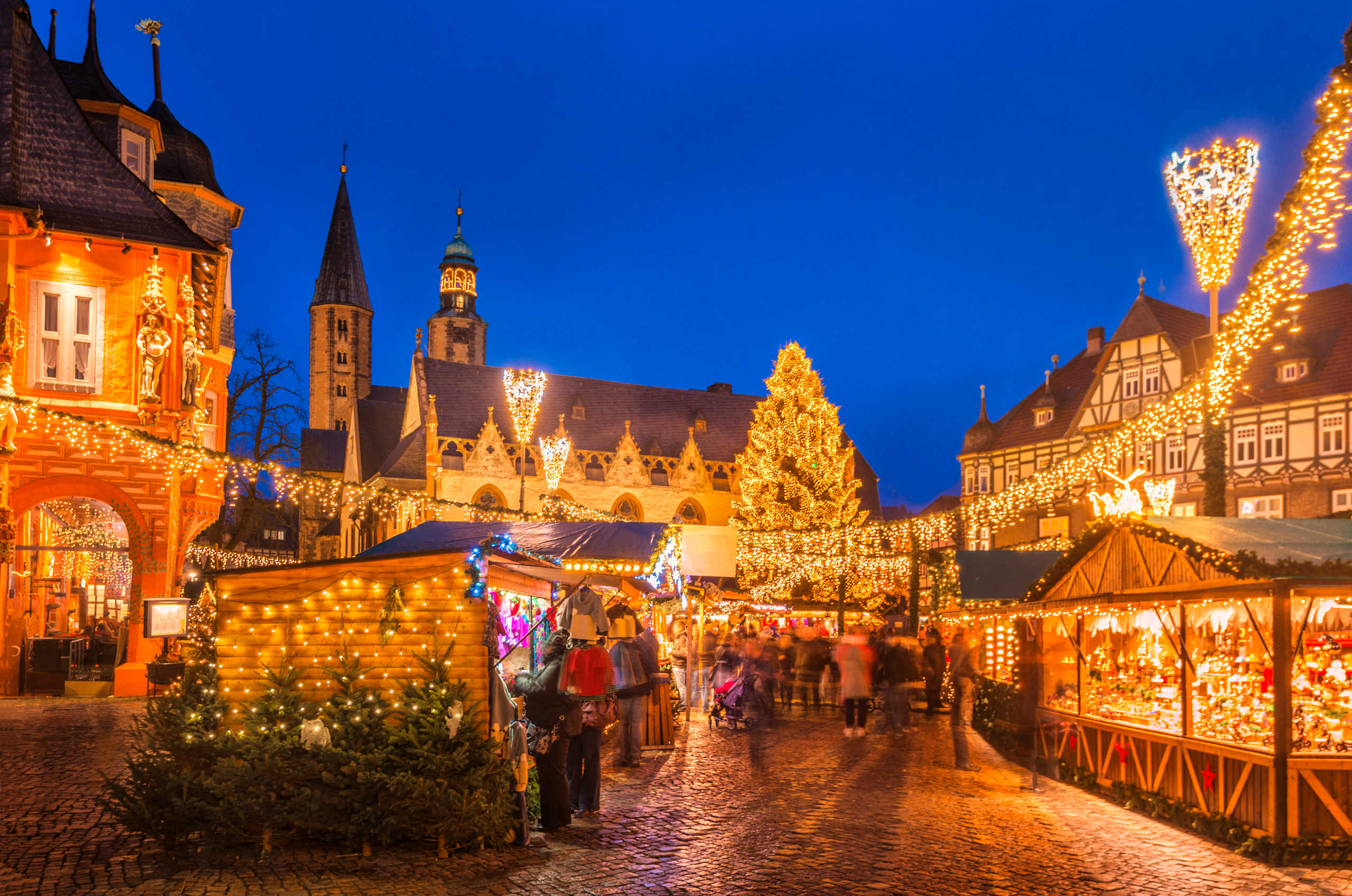 A bustling Christmas market with colorful stalls and twinkling lights, set against the backdrop of a historic town with a towering church spire.