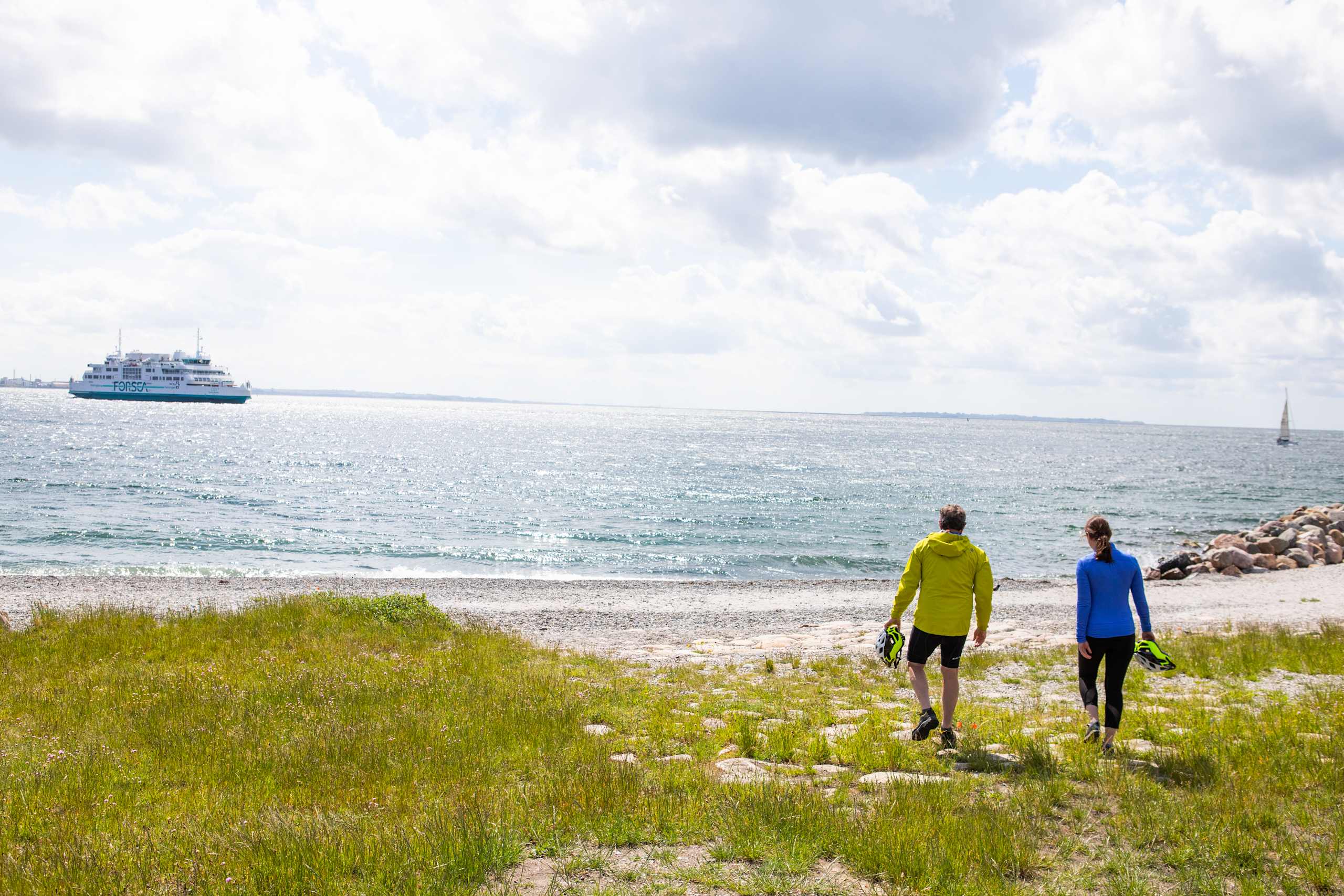 Two people in athletic wear walking along a grassy beach with a large cruise ship visible in the background on the calm, sparkling water.