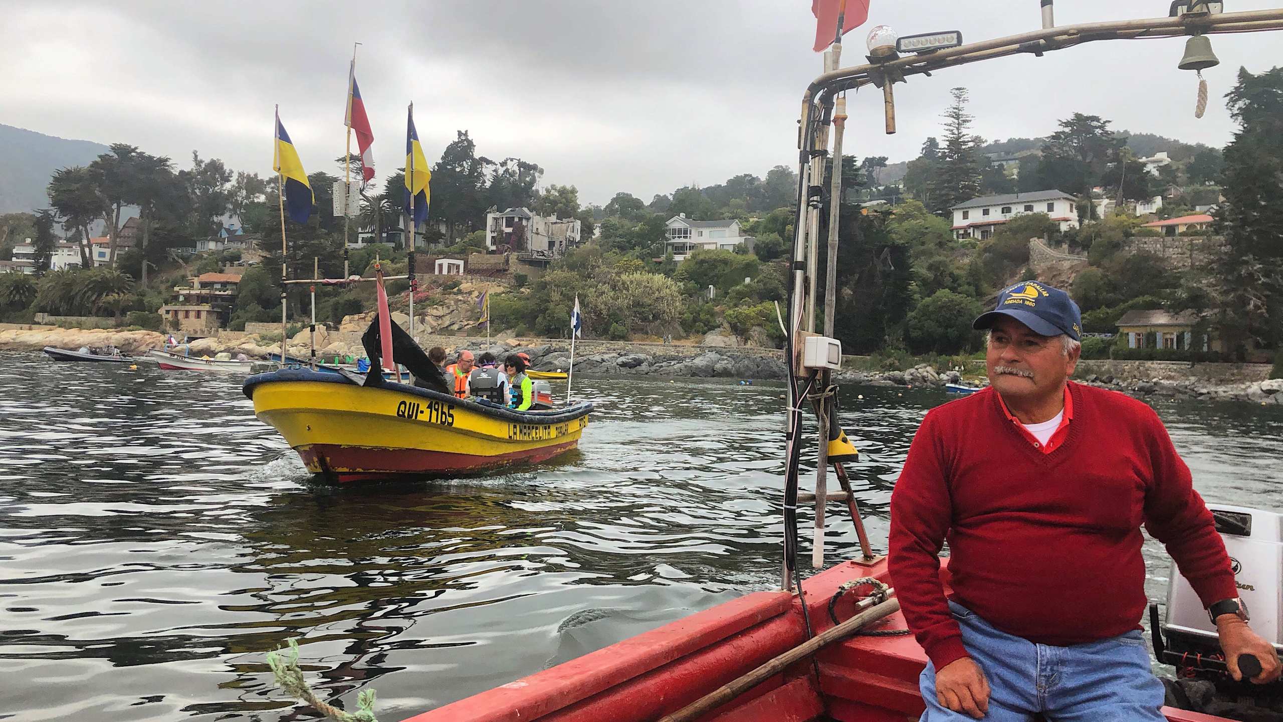 A colorful boat floats on a body of water surrounded by lush greenery and buildings, with a man in a red sweater standing on the shore.