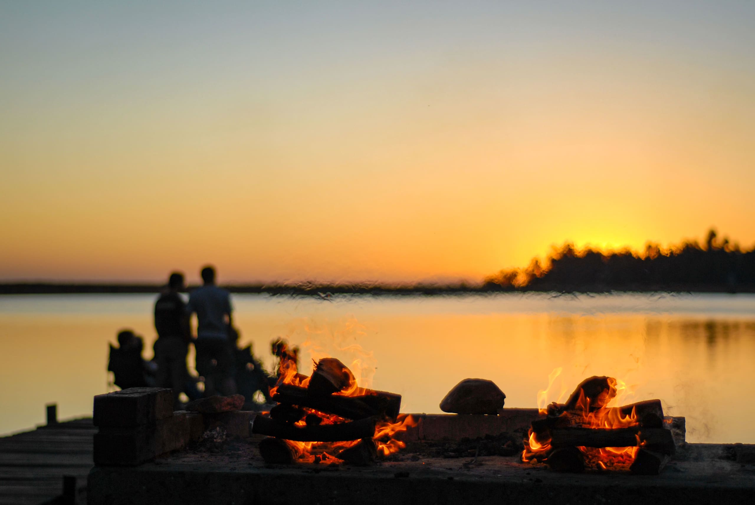 A serene lakeside scene with a campfire in the foreground and silhouettes of people against a vibrant sunset sky in the background.