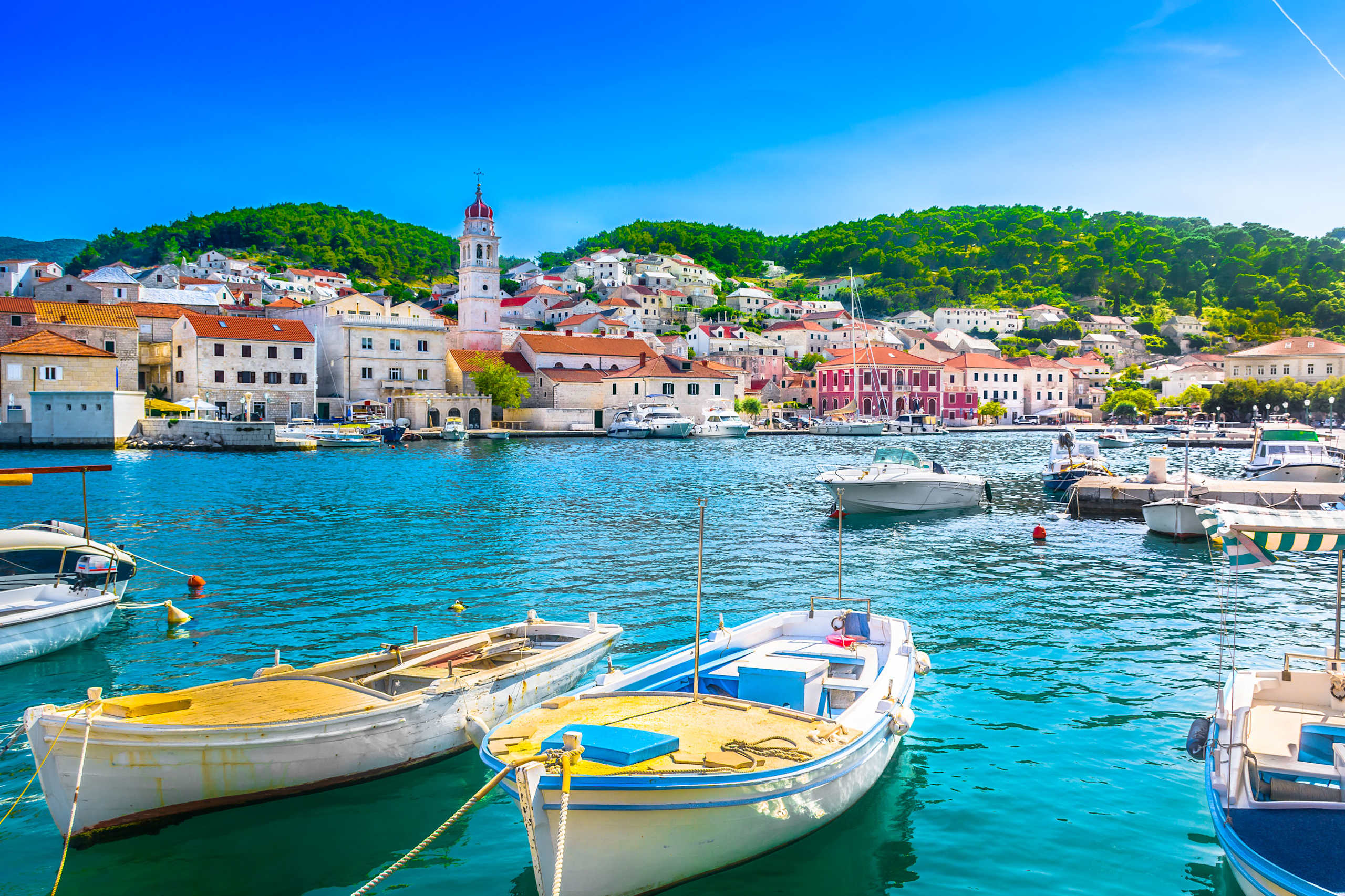 A picturesque harbor town with colorful boats floating on the turquoise waters, surrounded by lush green hills and a clear blue sky.