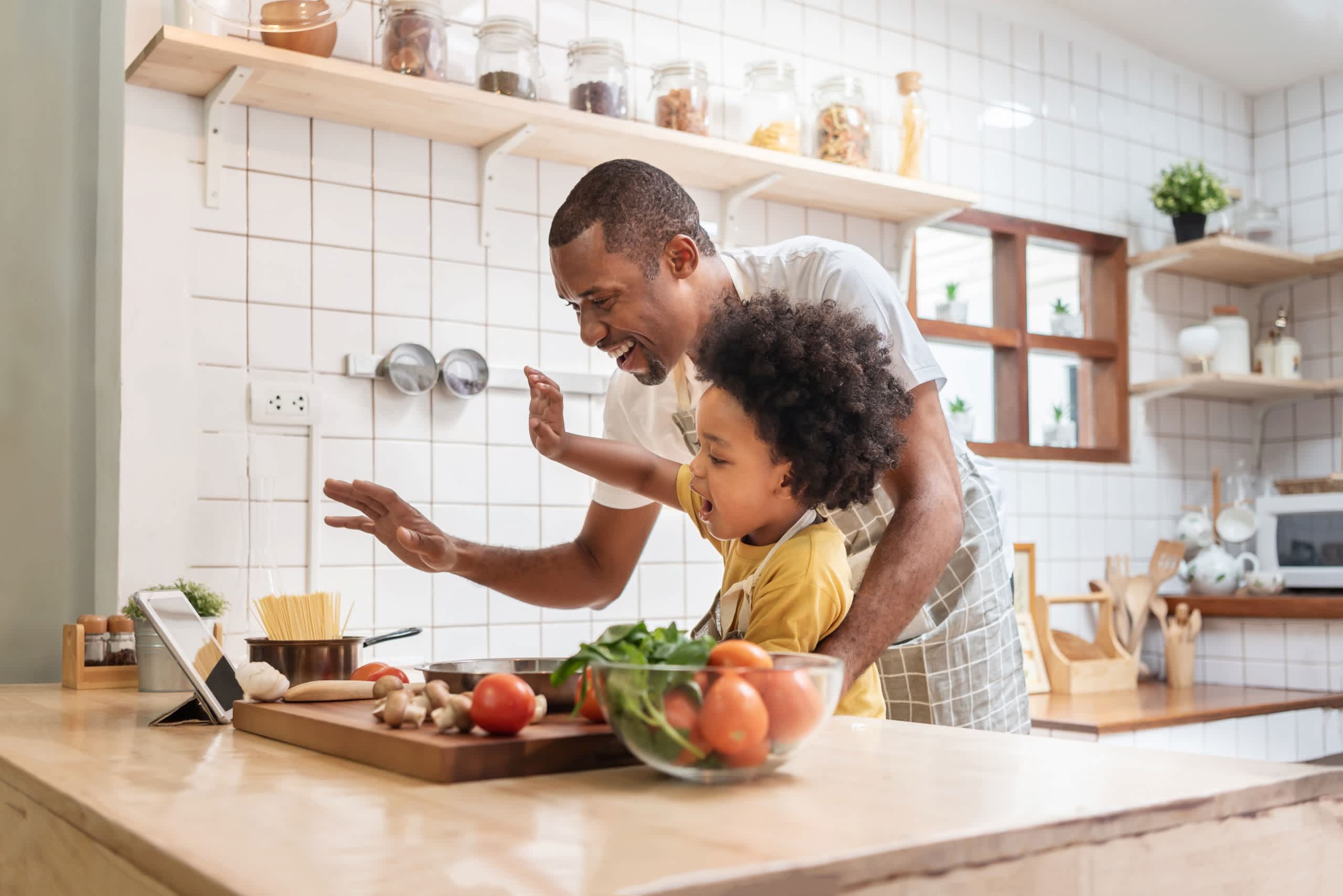 A man and a young child are standing together in a kitchen, preparing a meal with fresh produce on the counter in front of them.