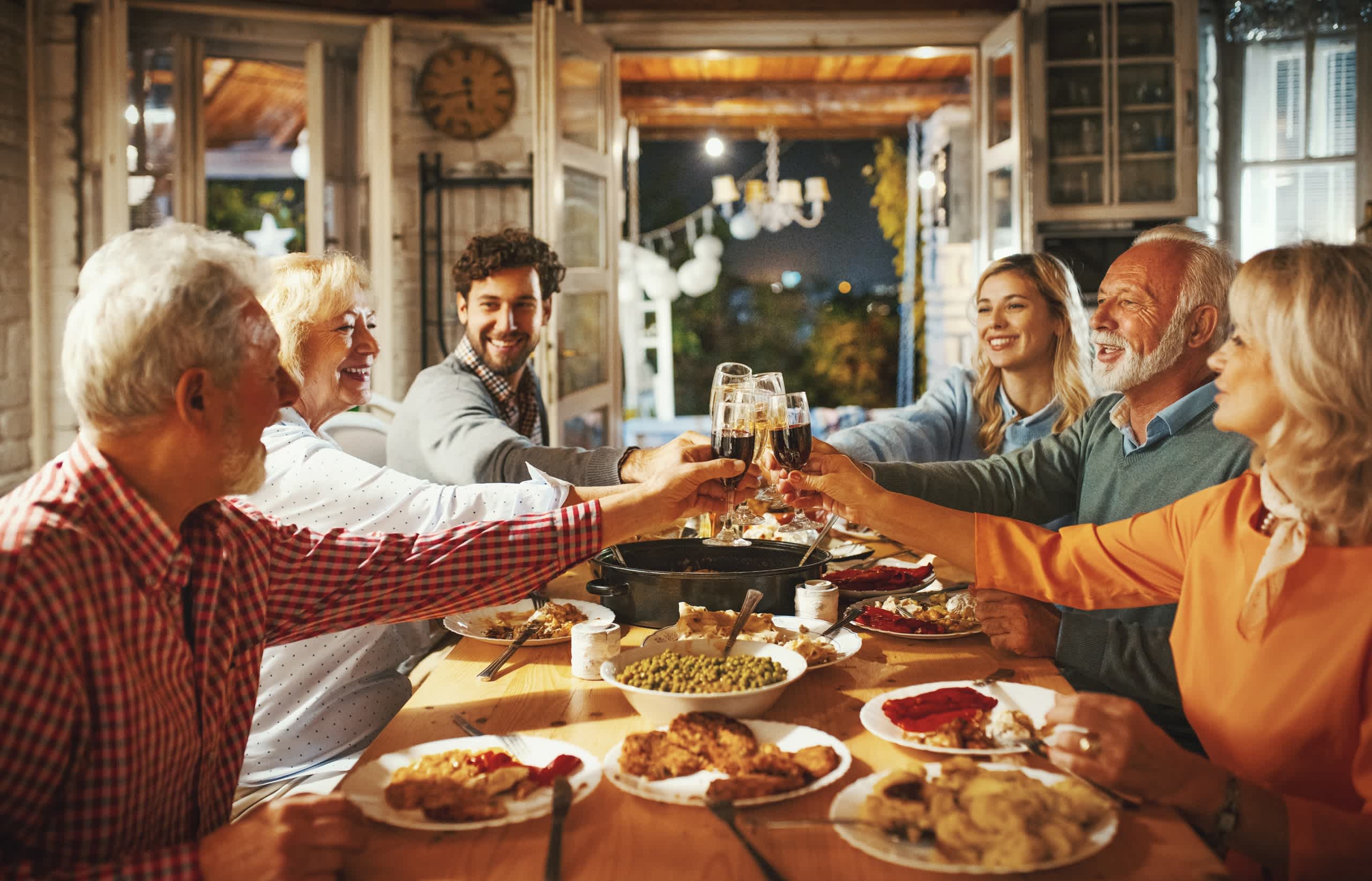 A group of people, both young and old, are gathered around a table enjoying a festive meal together, with various dishes and drinks visible in the foreground, and a cozy, rustic interior setting in the background.