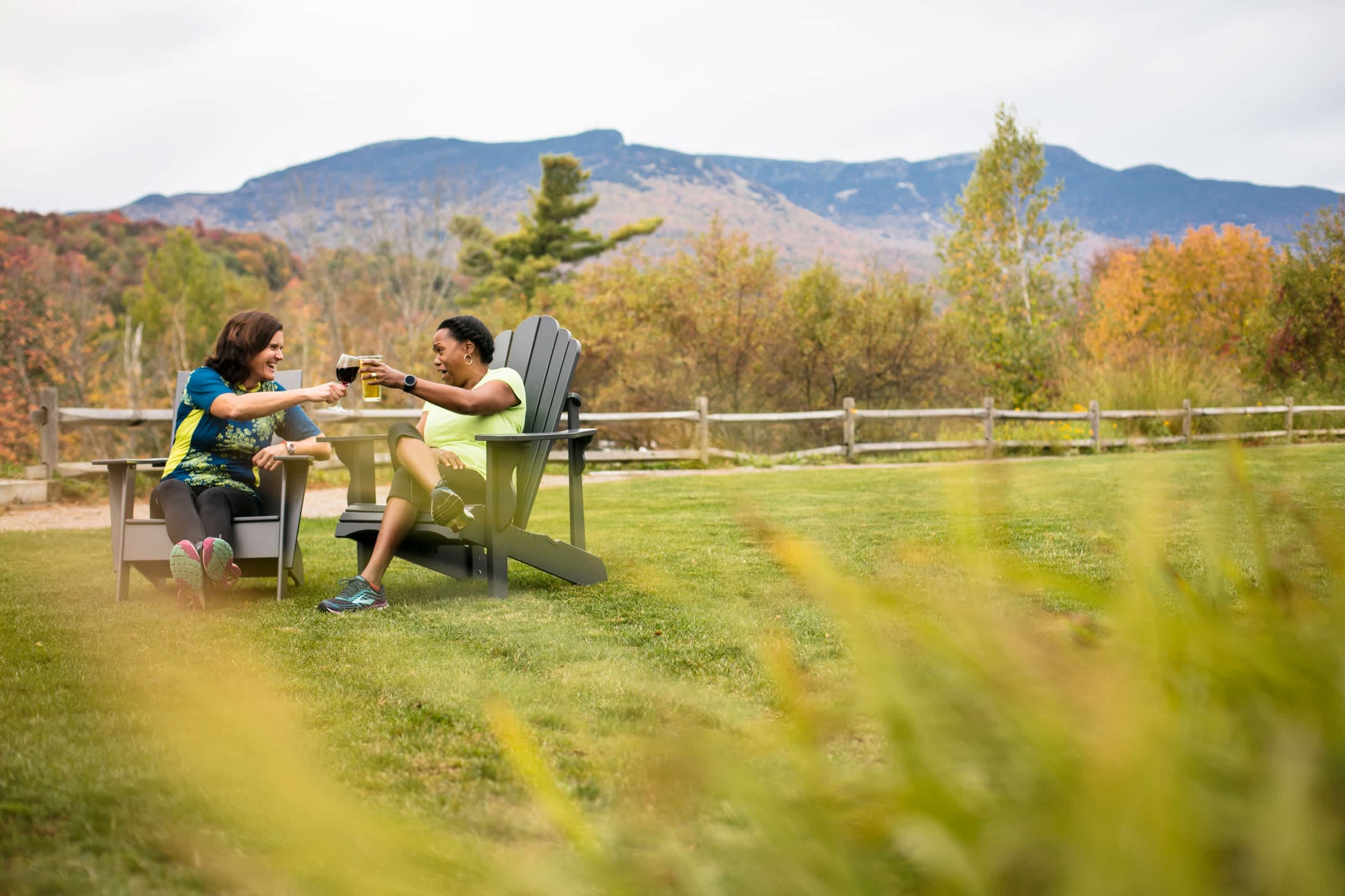 Two people, a man and a woman, are sitting on Adirondack chairs in a grassy field, enjoying drinks and taking in the scenic mountain landscape in the background.