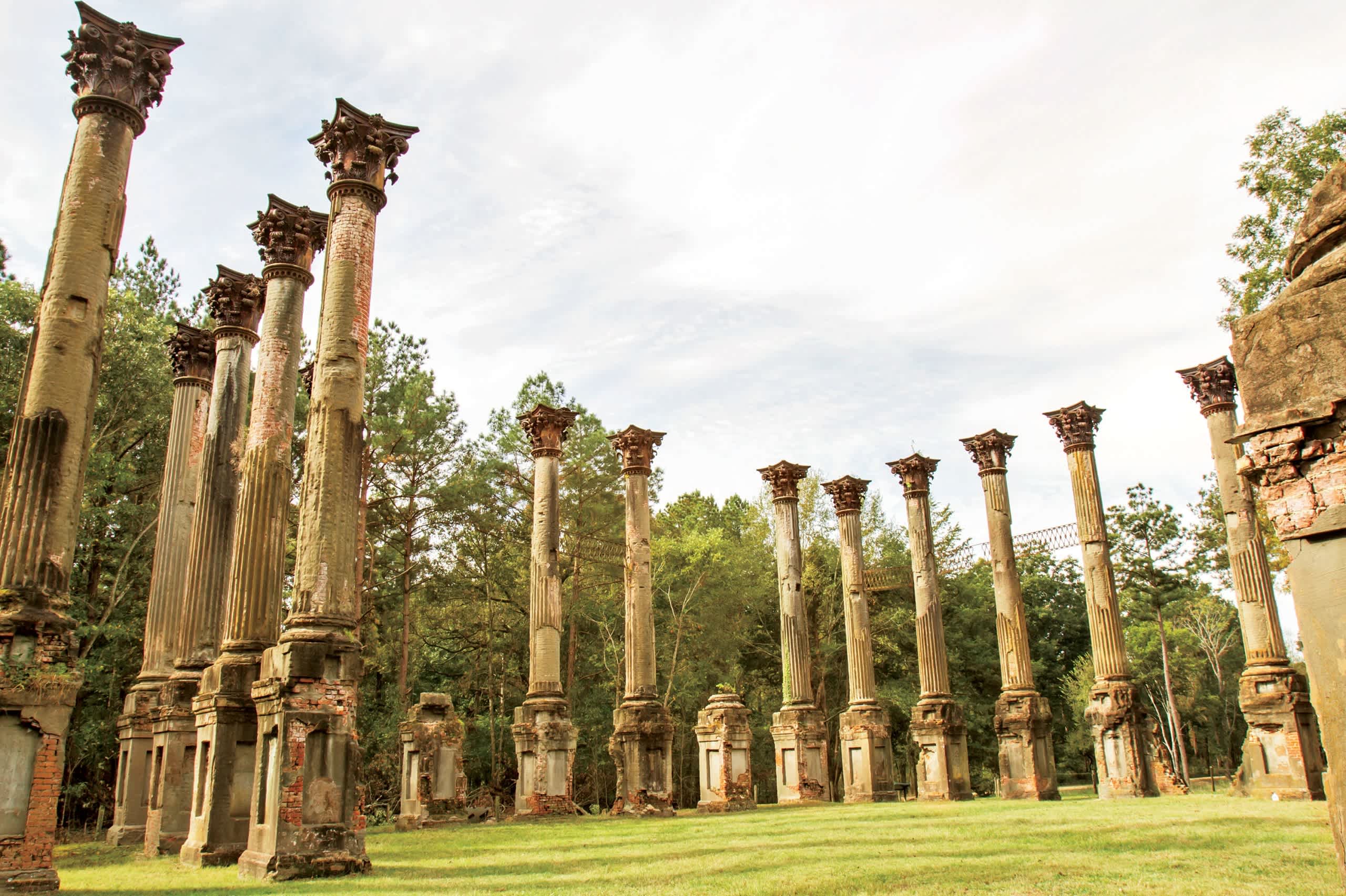 Rows of ornate stone columns stand tall in a grassy field, surrounded by lush greenery and a cloudy sky.