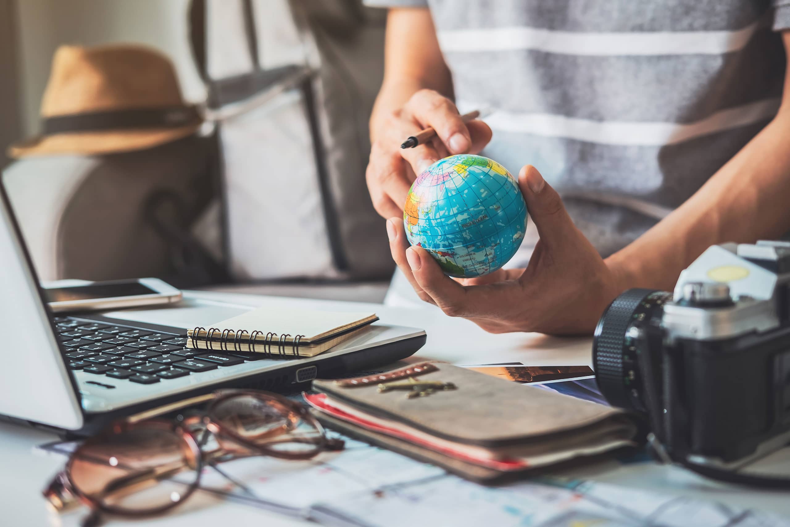 The image shows a person's hands holding a small globe or model of the Earth, surrounded by various office supplies and a laptop on a desk.