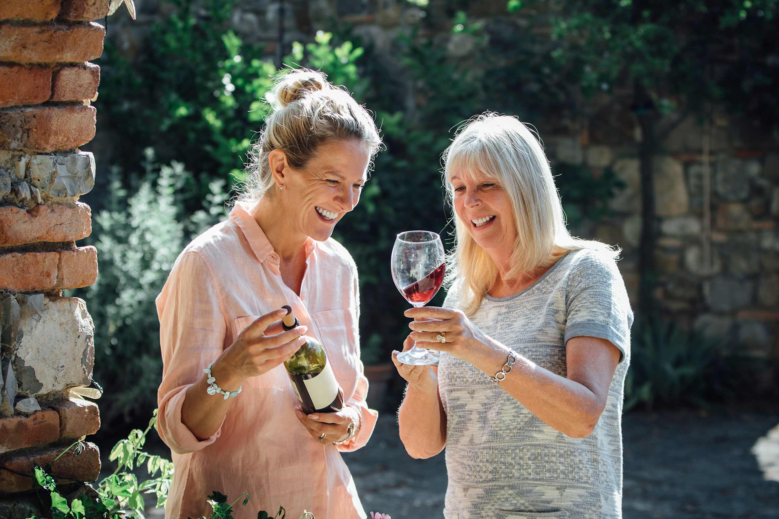 Two women, one with blonde hair and the other with gray hair, are standing in a garden setting and enjoying glasses of wine together.