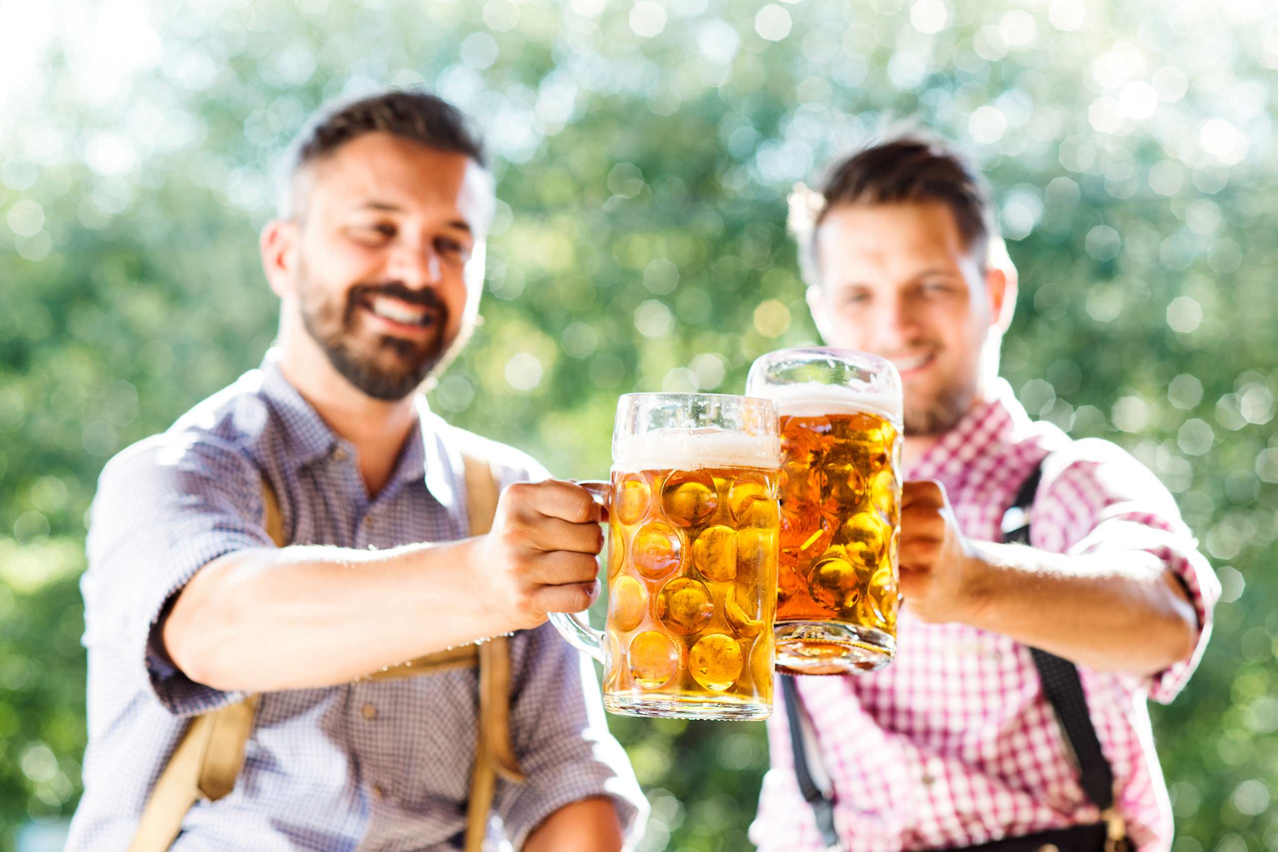 Two men in casual attire are holding up large glasses of beer, with a blurred natural background of greenery visible behind them.