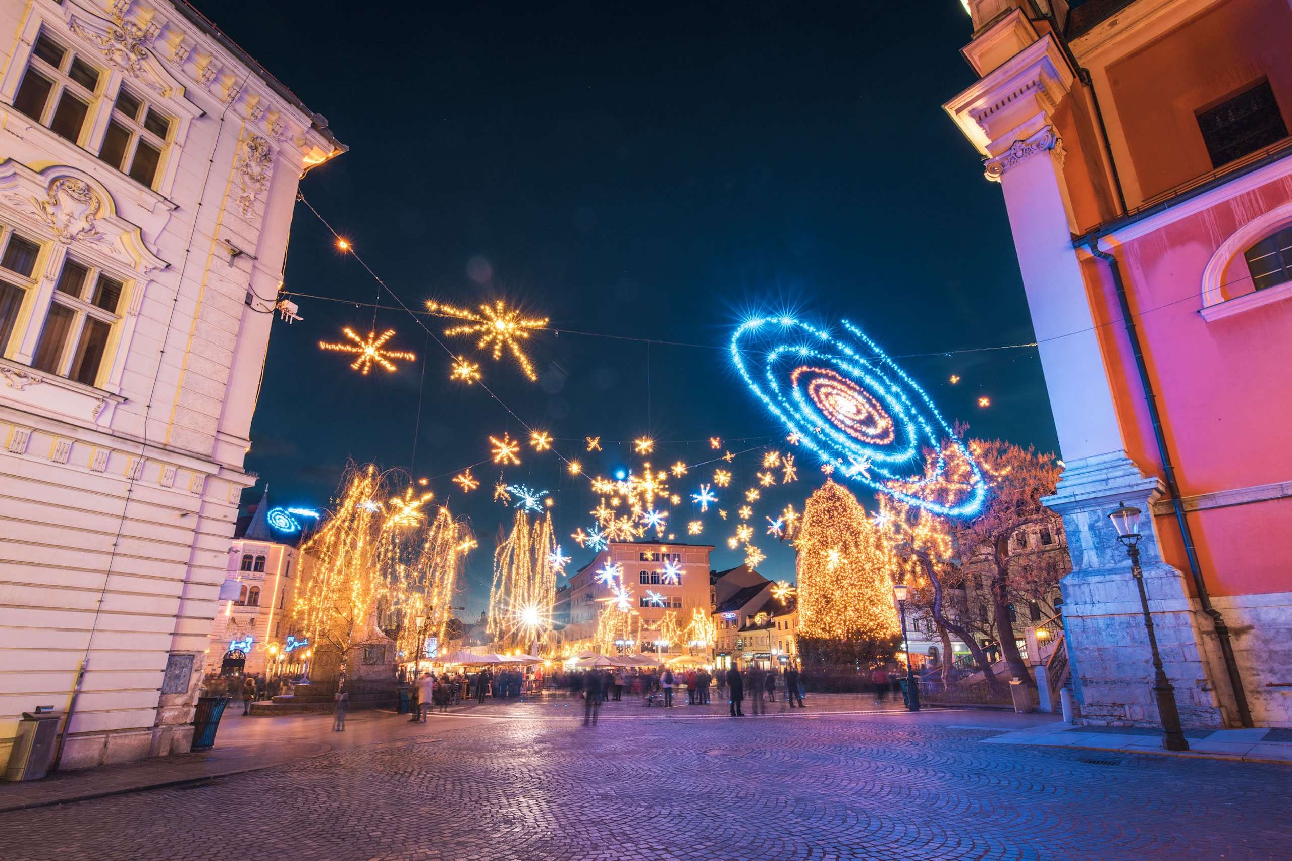 A vibrant and festive nighttime scene with colorful illuminated decorations, including large star-shaped lights and a large, intricate light display in the background, set against the backdrop of historic buildings in a city square.