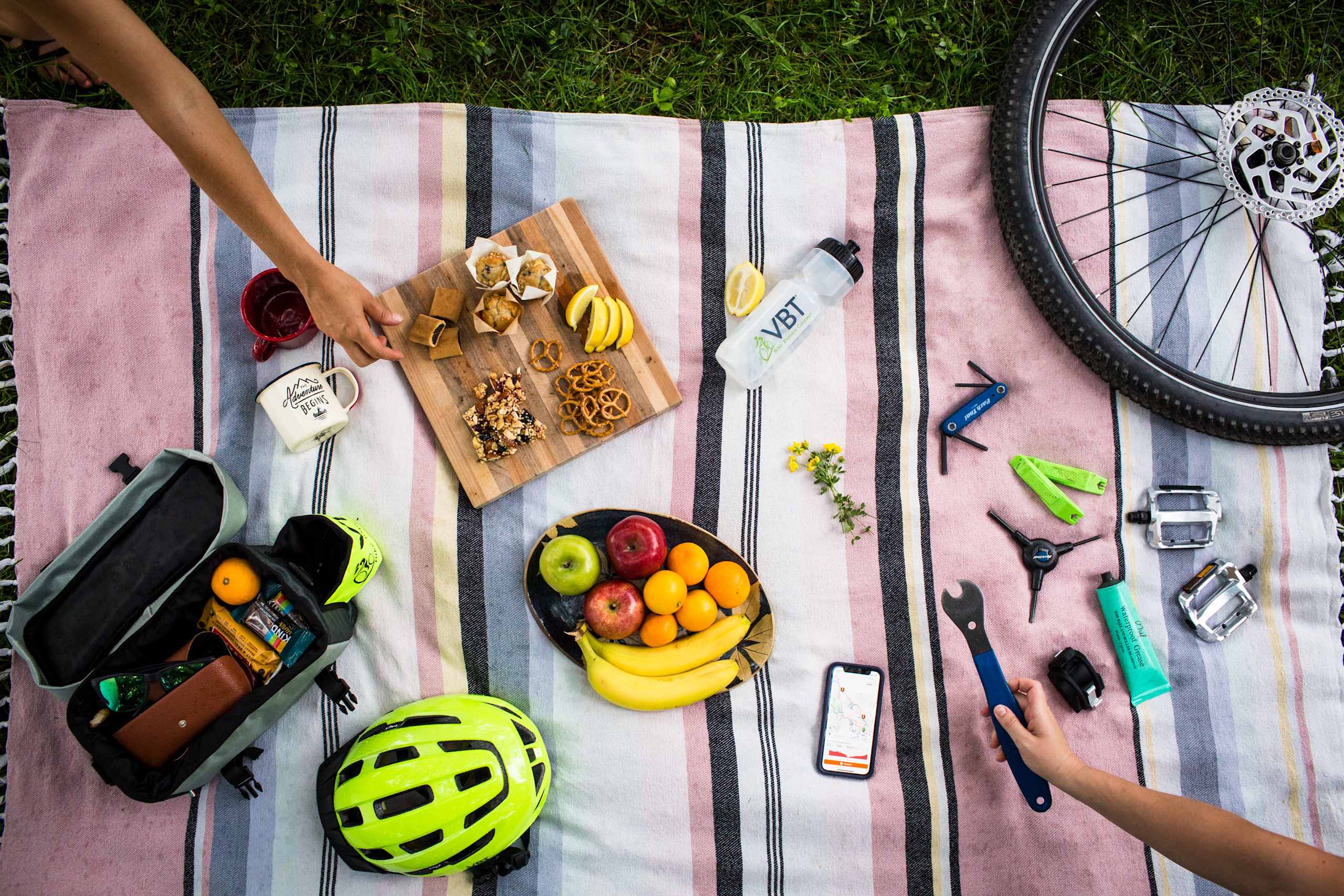 A picnic blanket laid out on the grass, filled with various items such as fruits, tools, and a bicycle wheel, creating a vibrant and organized outdoor scene.