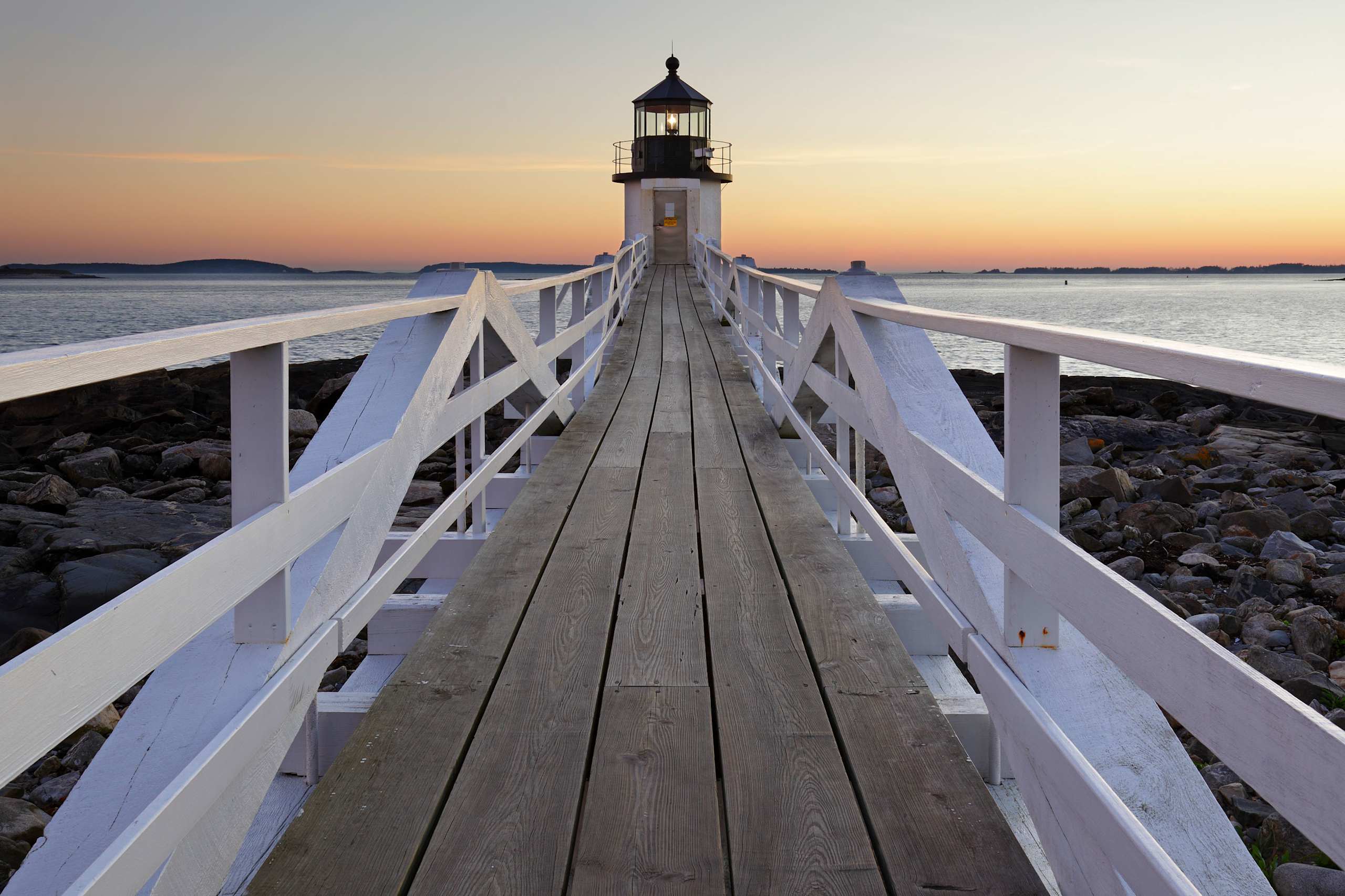 A wooden pier leads to a lighthouse standing tall against a vibrant sunset sky, with the calm waters of the bay stretching out in the background.