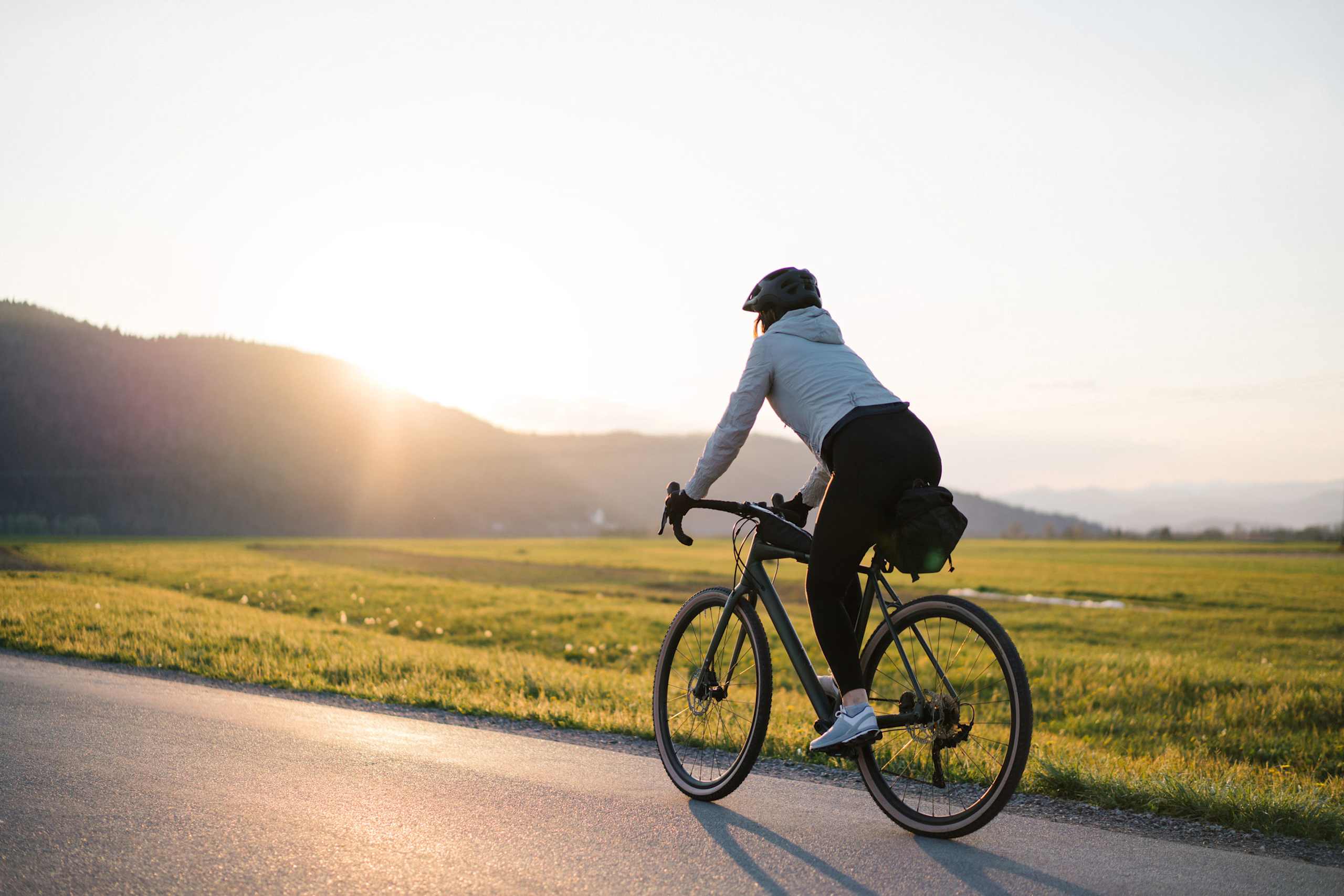 A person on a bicycle rides through a scenic countryside landscape with mountains in the background and a golden field in the foreground.