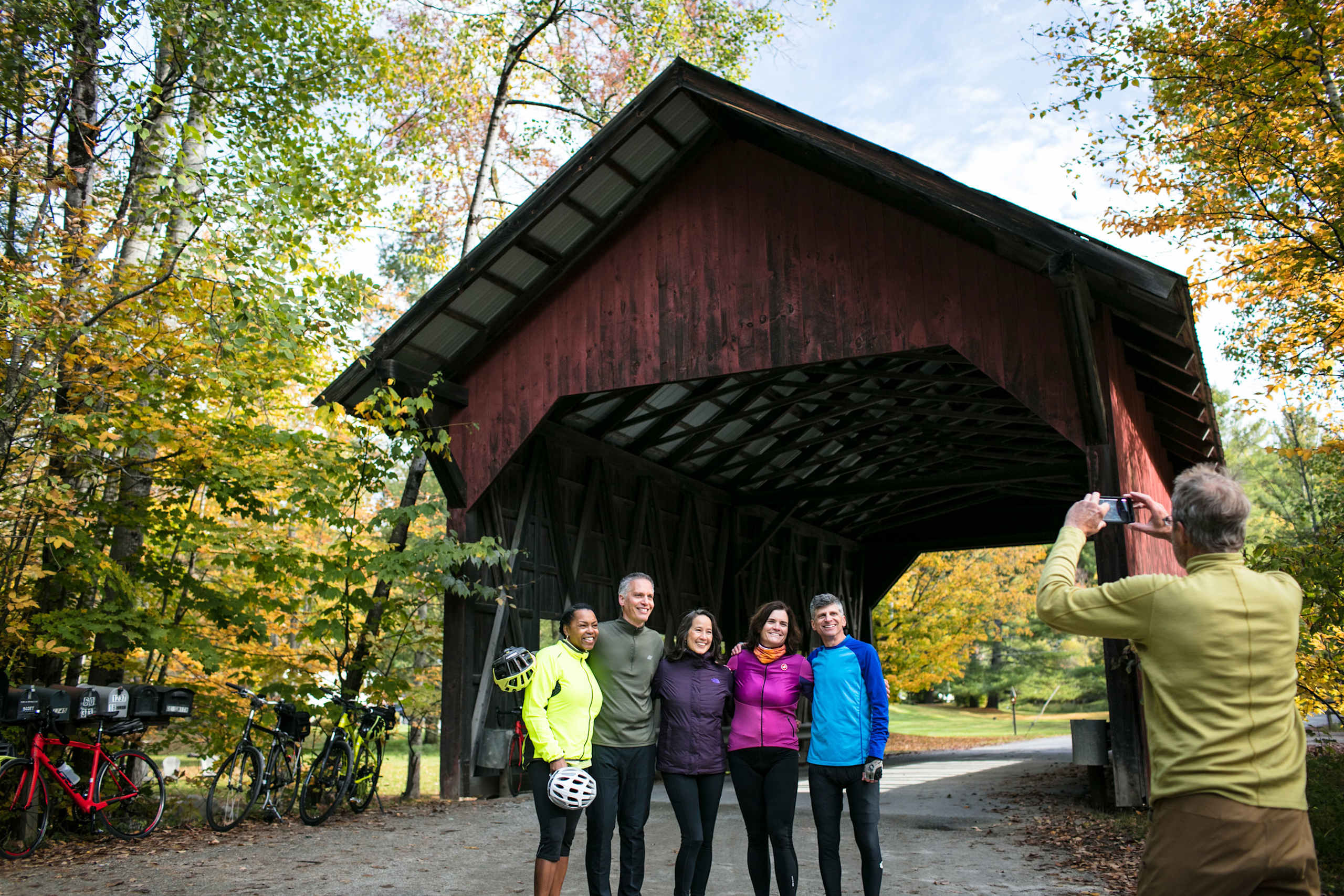 A group of people standing in front of a red wooden covered bridge, surrounded by a colorful autumn landscape with trees and a paved path.