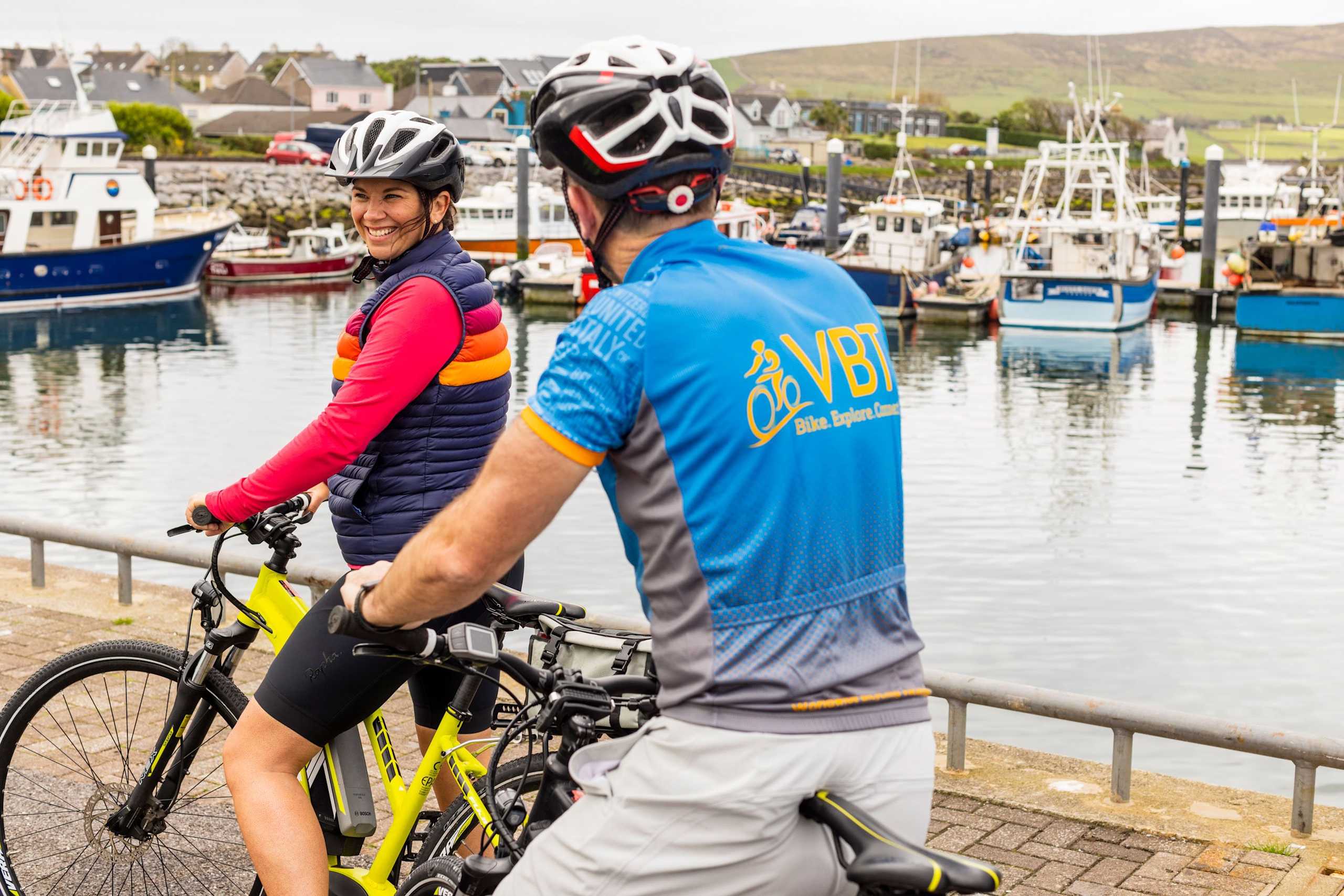 Two cyclists, one wearing a red jacket and the other a blue jersey, stand on a dock overlooking a harbor with boats in the background.