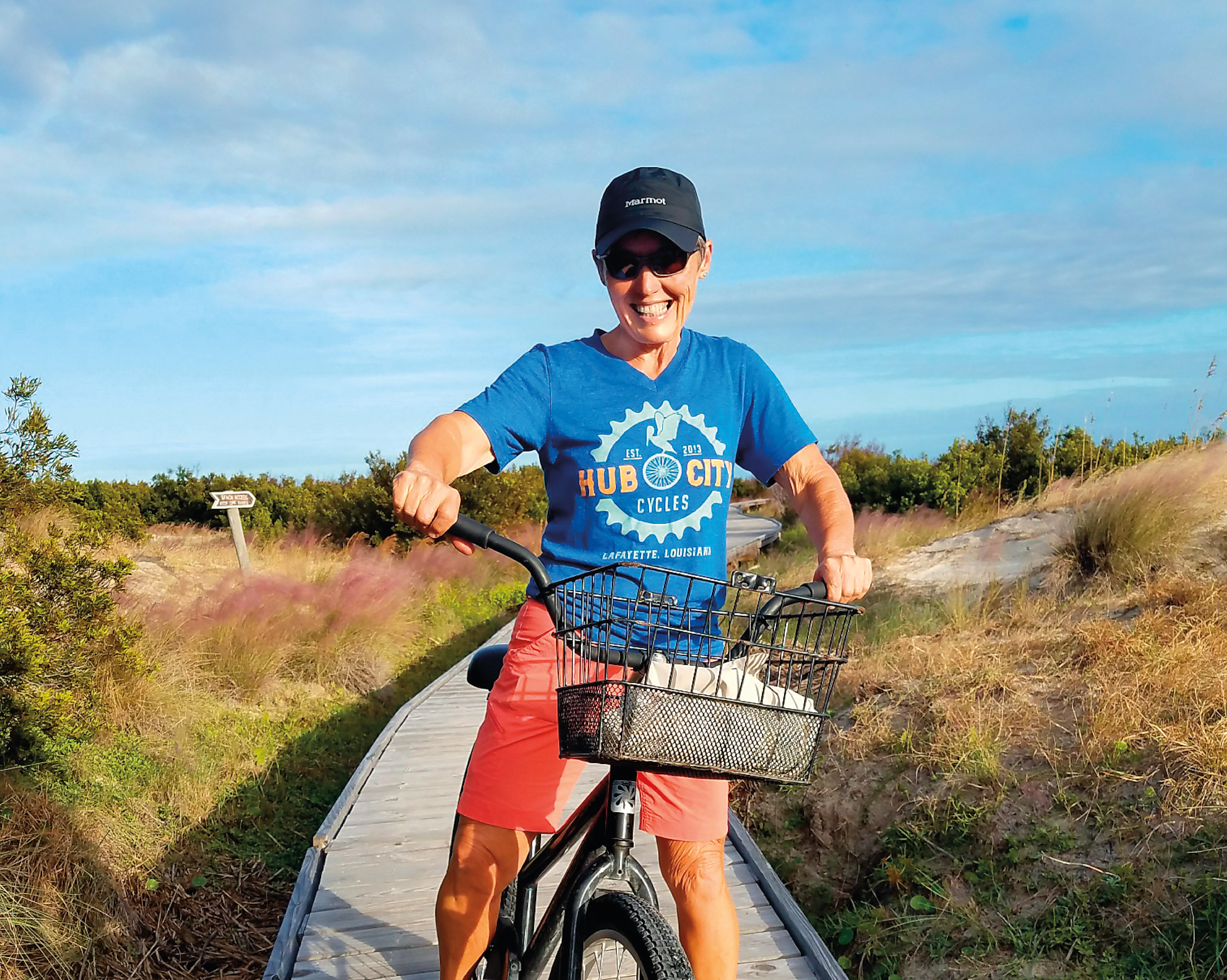 A person wearing a blue t-shirt and a baseball cap is riding a bicycle on a wooden walkway surrounded by natural scenery, including trees and a clear sky.