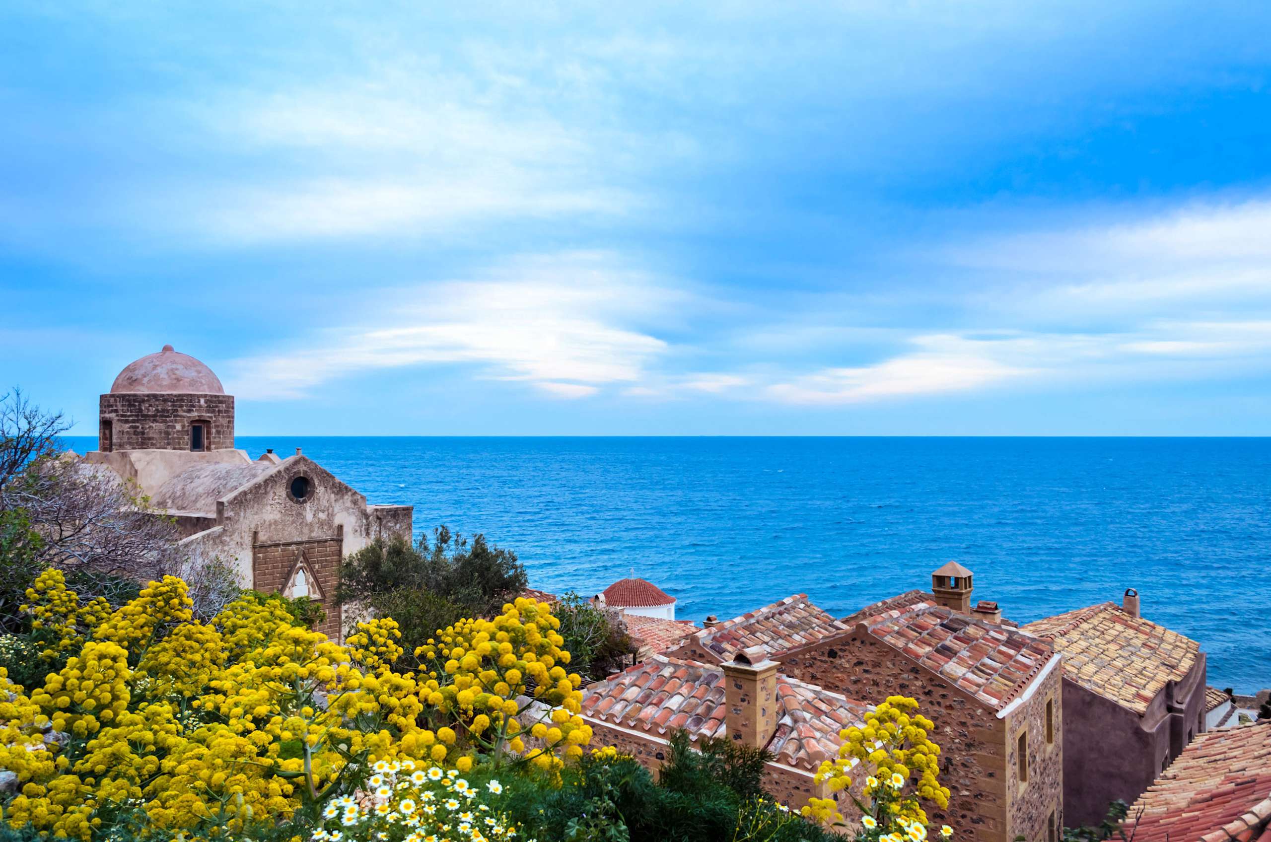 A picturesque coastal town with a historic church or building nestled among vibrant yellow flowers, set against a backdrop of a serene blue ocean and sky.