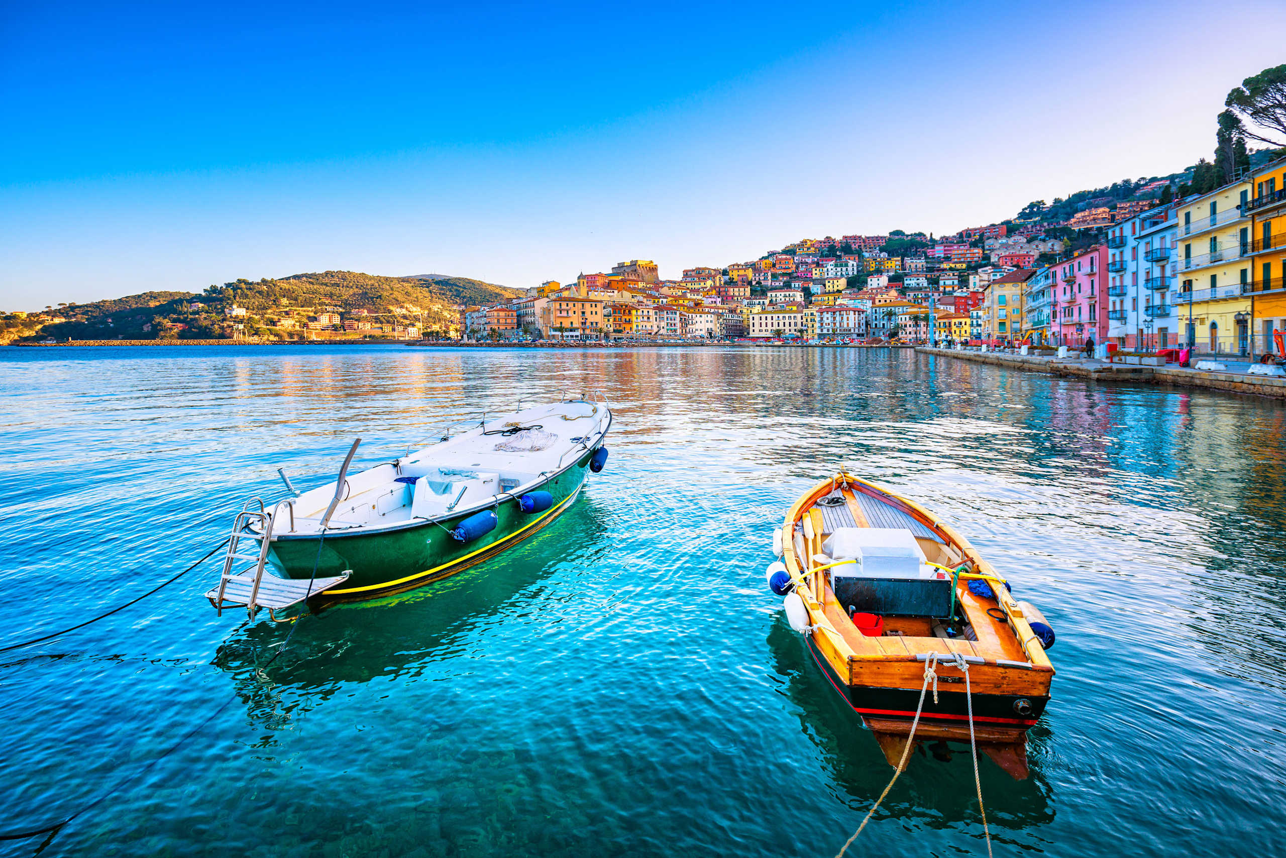 A picturesque harbor with colorful buildings lining the shore, reflected in the calm, turquoise waters, with two small boats floating in the foreground.